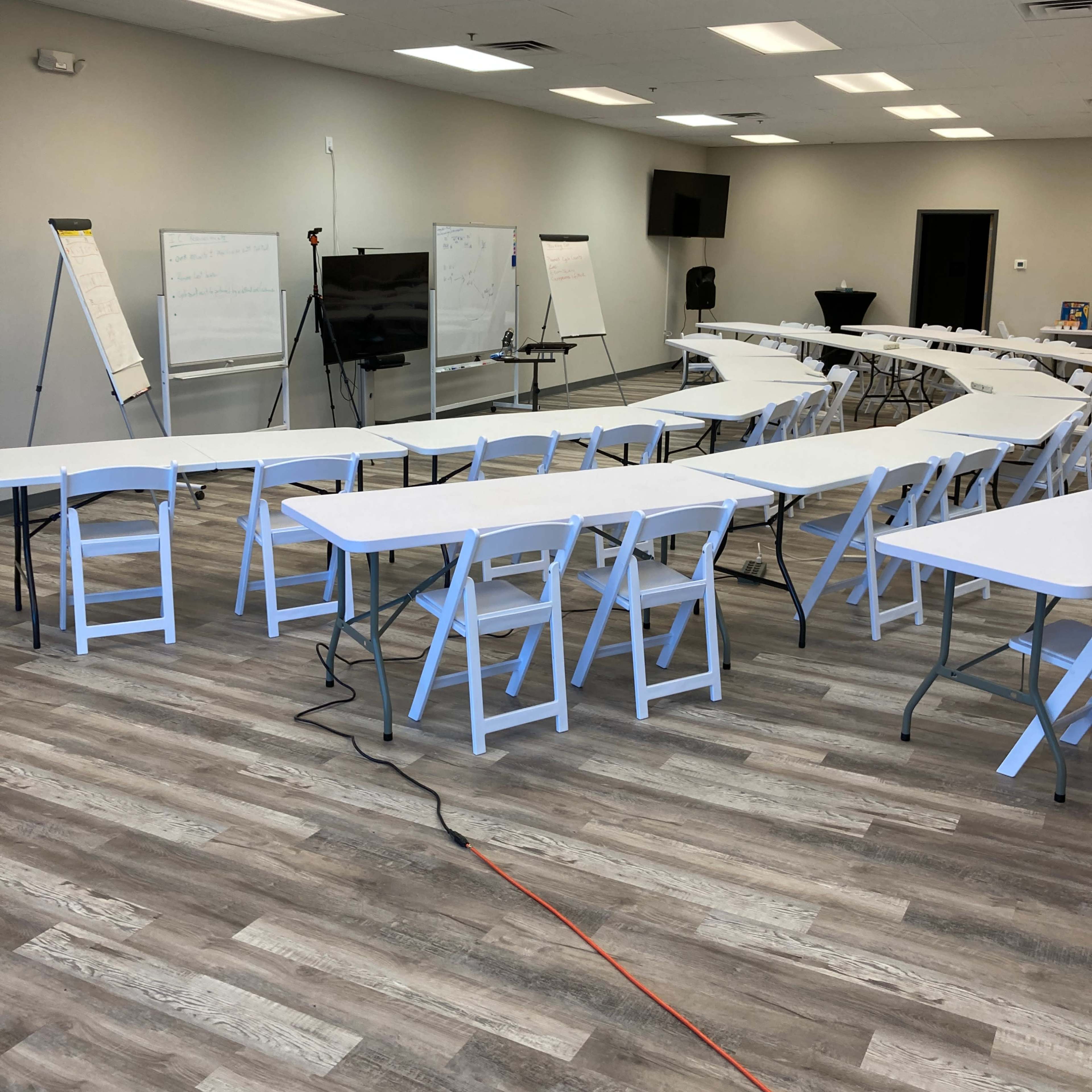 A spacious training room contains several white tables arranged in rows, along with flip charts and a presentation screen at the front.