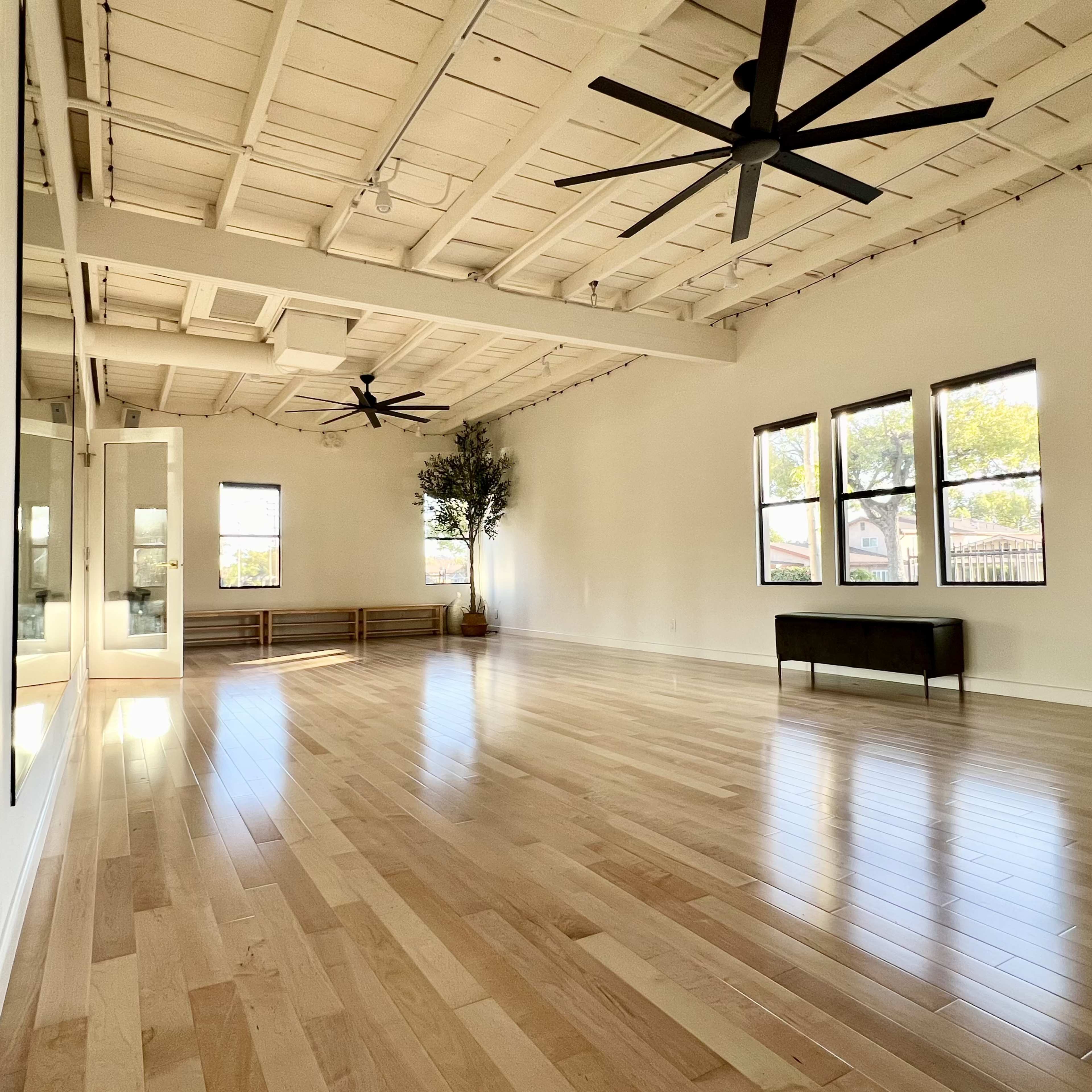 A spacious, well-lit room with hardwood flooring, two ceiling fans, and large windows.