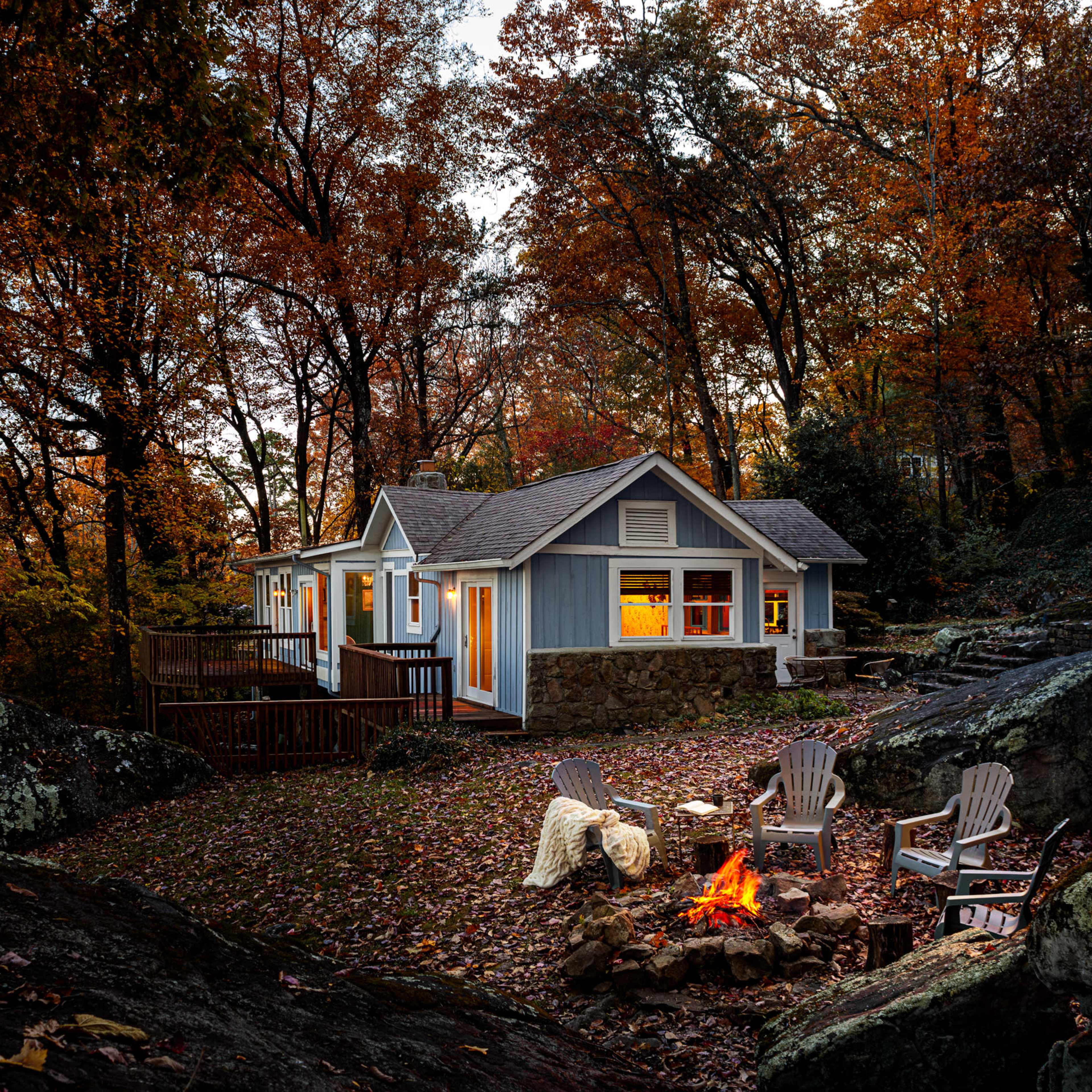 A cozy house with warm lights sits surrounded by autumn trees, with a fire pit and chairs arranged in the foreground.