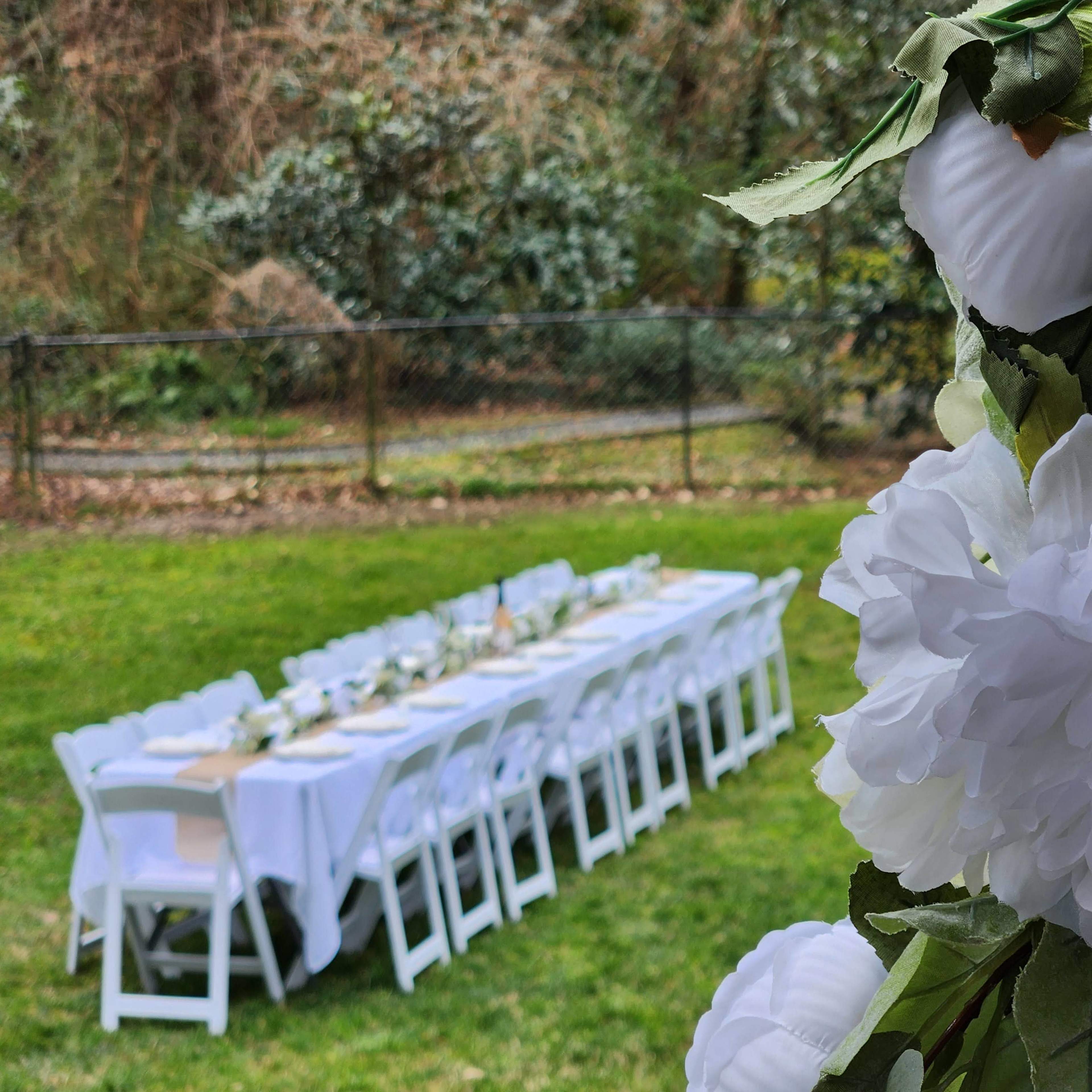 A long table is set with white tablecloths and chairs in a grassy backyard, adorned with white flowers on one side.