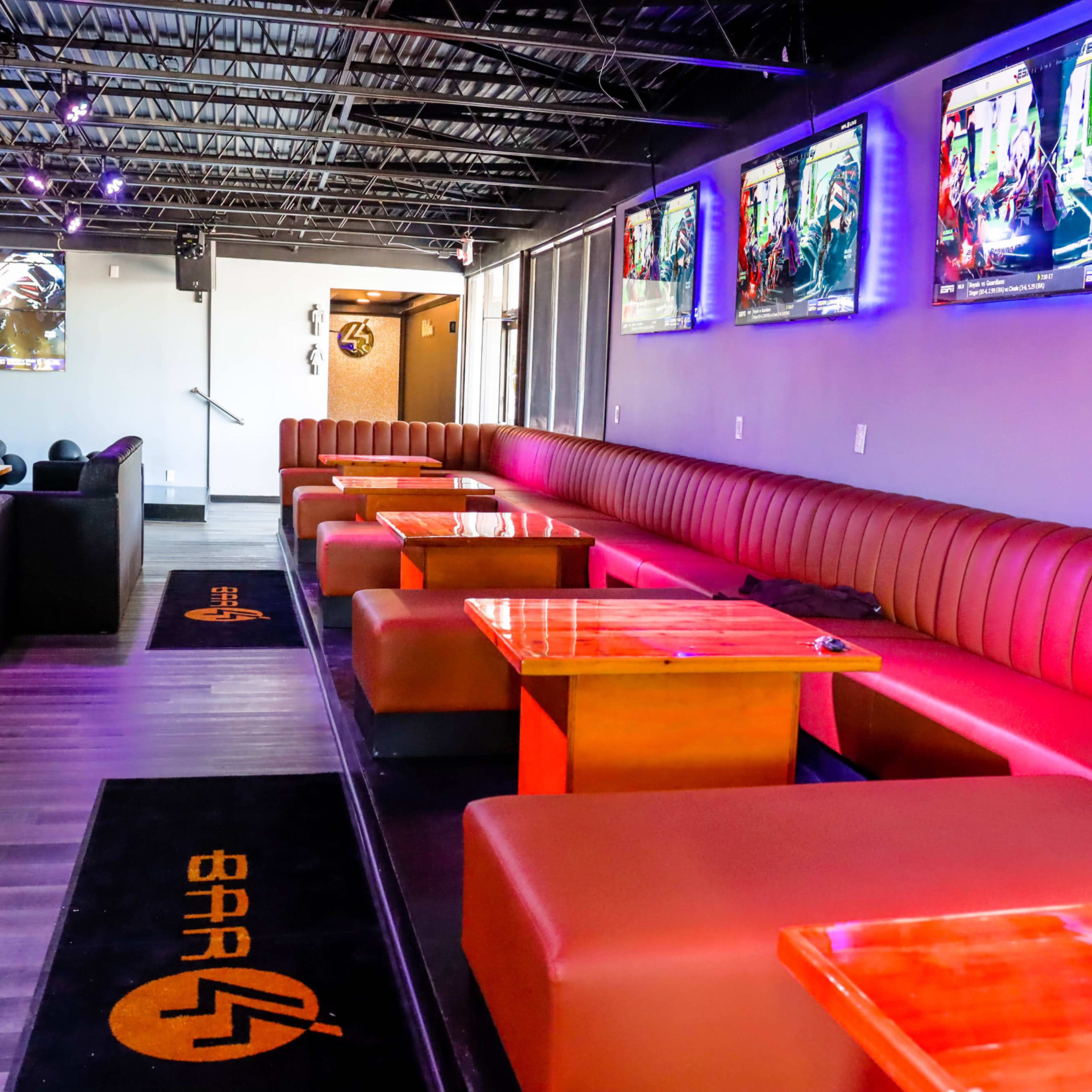 A modern lounge area with plush seating, wooden tables, and multiple screens displaying colorful visuals in a well-lit space.