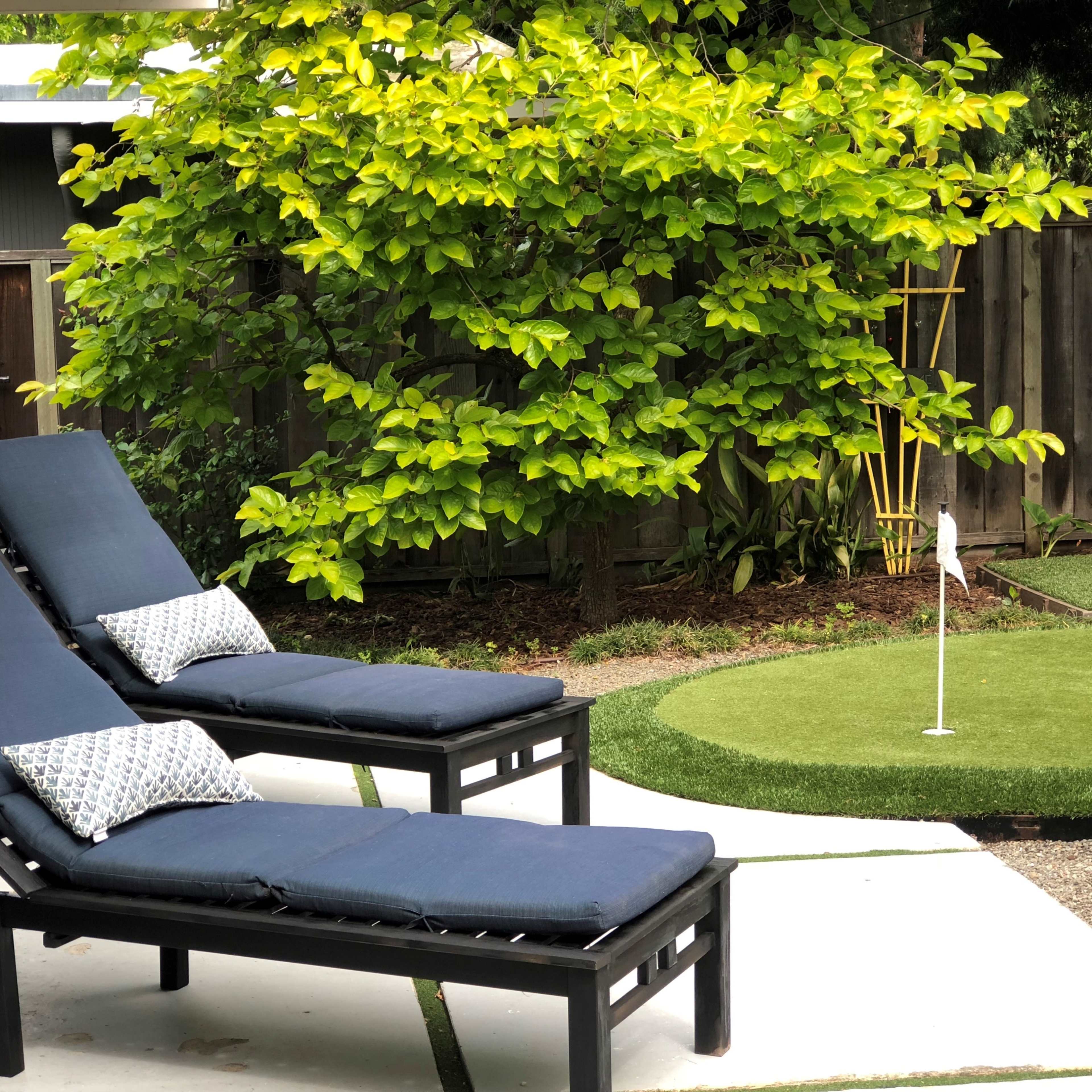 Two black lounge chairs with cushions beside a small putting green surrounded by greenery in a backyard.
