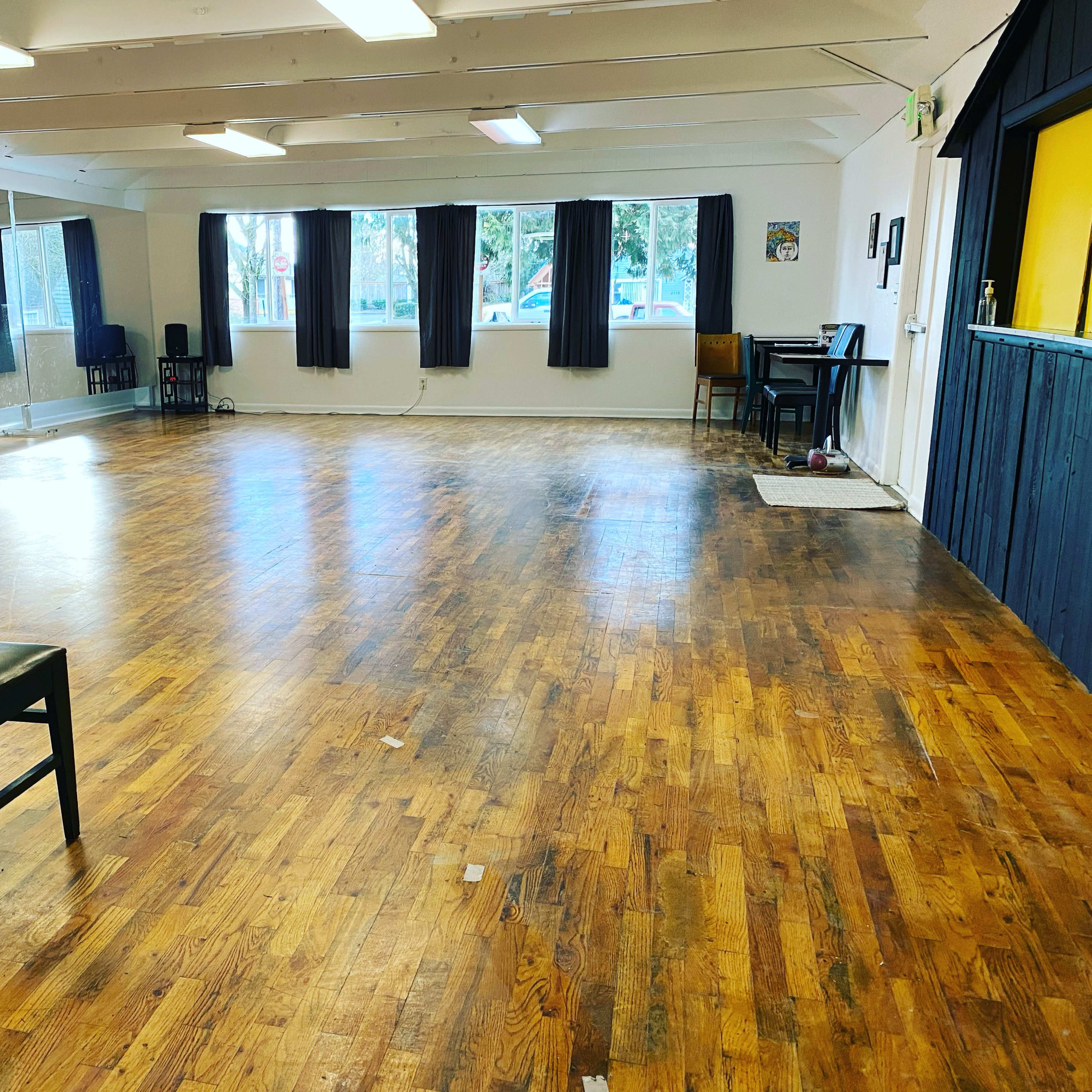 A spacious dance studio with wooden floors, mirrors on one wall, and several windows allowing natural light.
