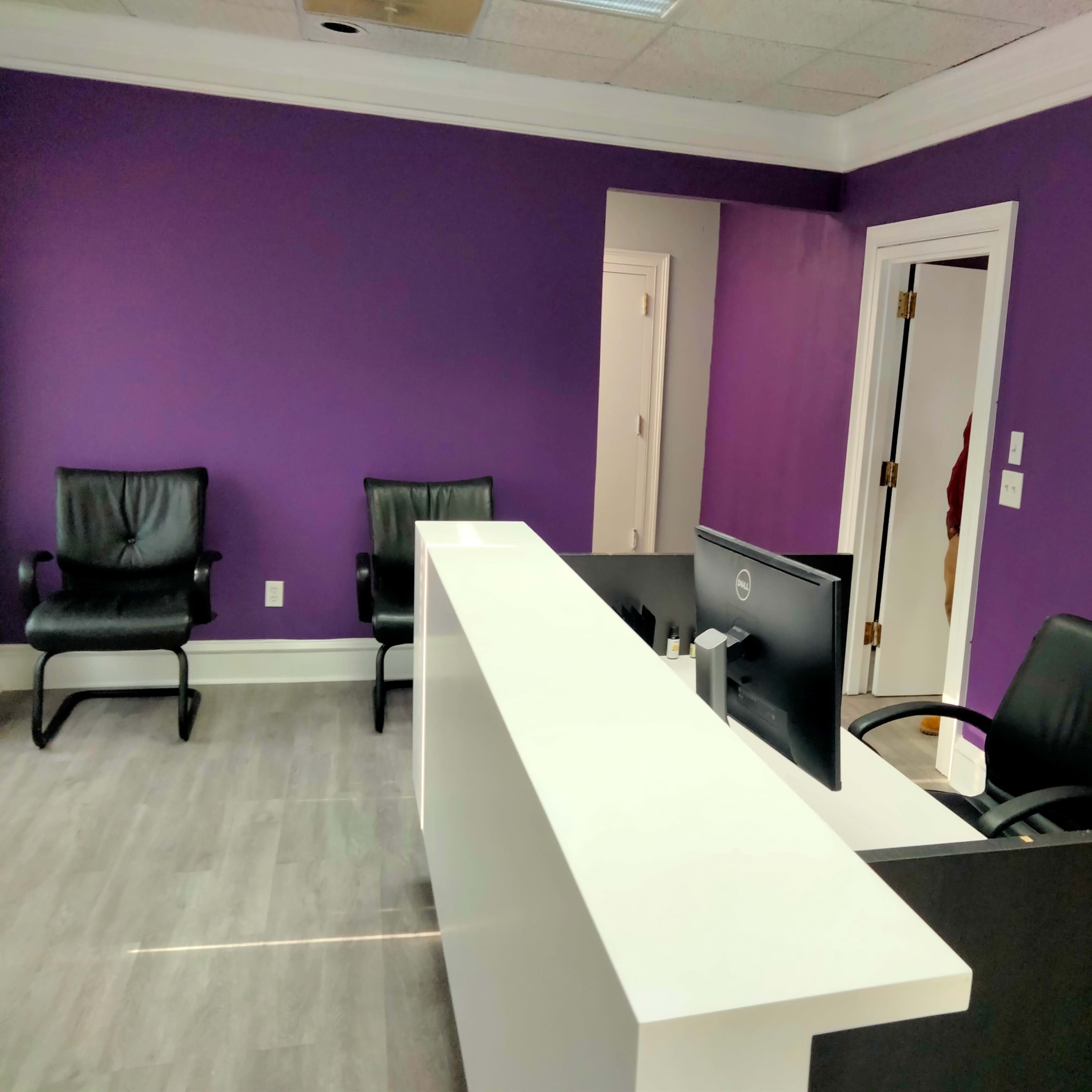 A modern office reception area with a white front desk, a computer monitor, and two black chairs against a purple wall.