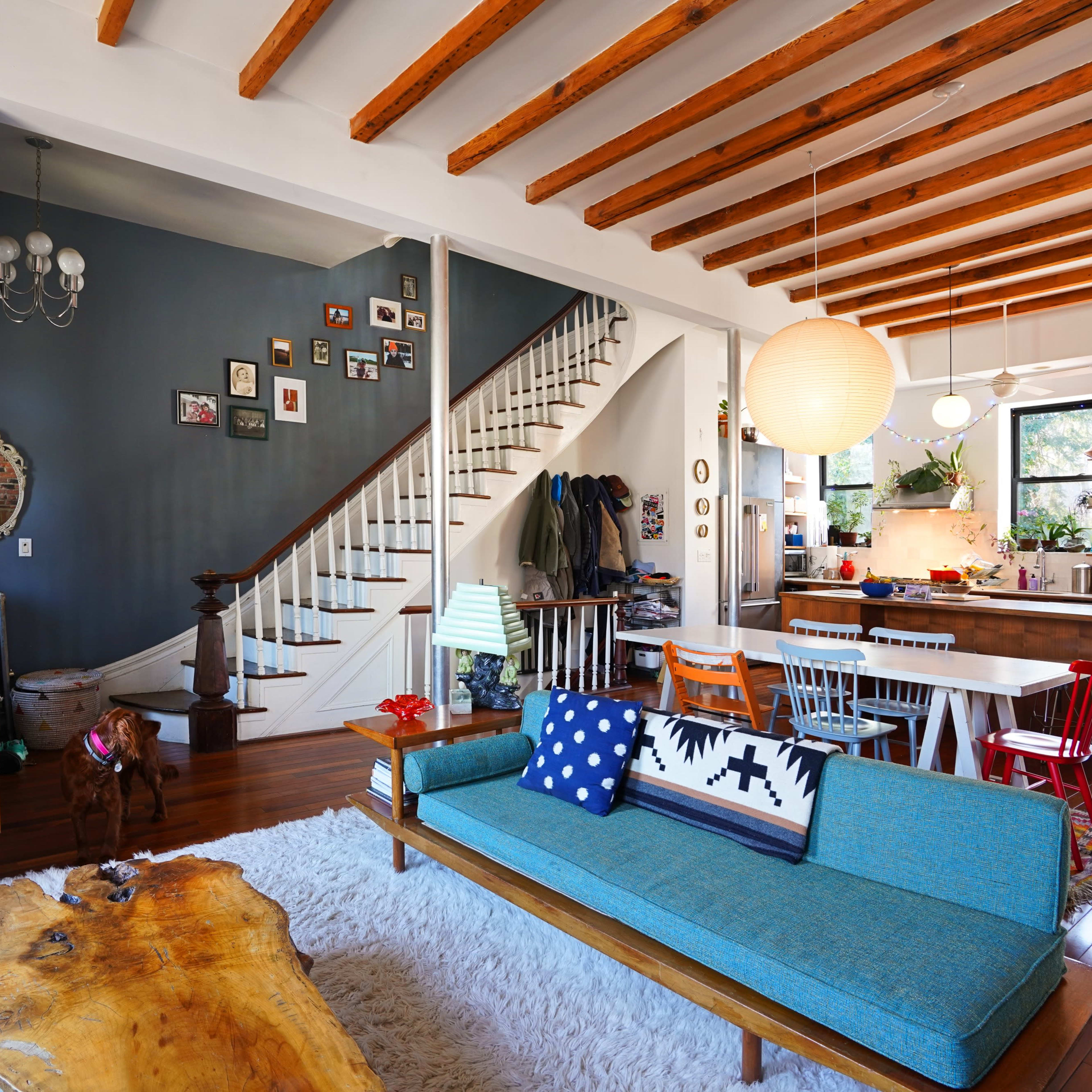 A bright living space with wooden beams, a blue sofa, and an open layout that connects to a staircase and kitchen area.