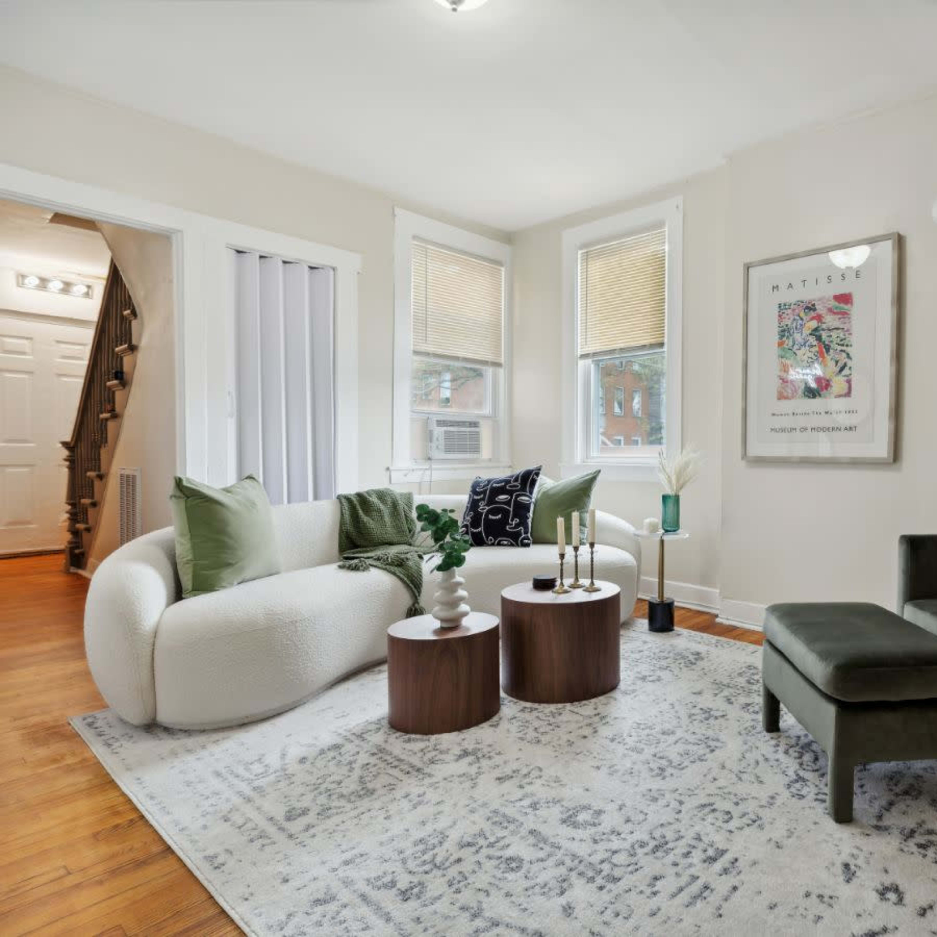 A modern living room furnished with a white circular sofa, a green armchair, two wooden side tables with candles, and a large area rug.