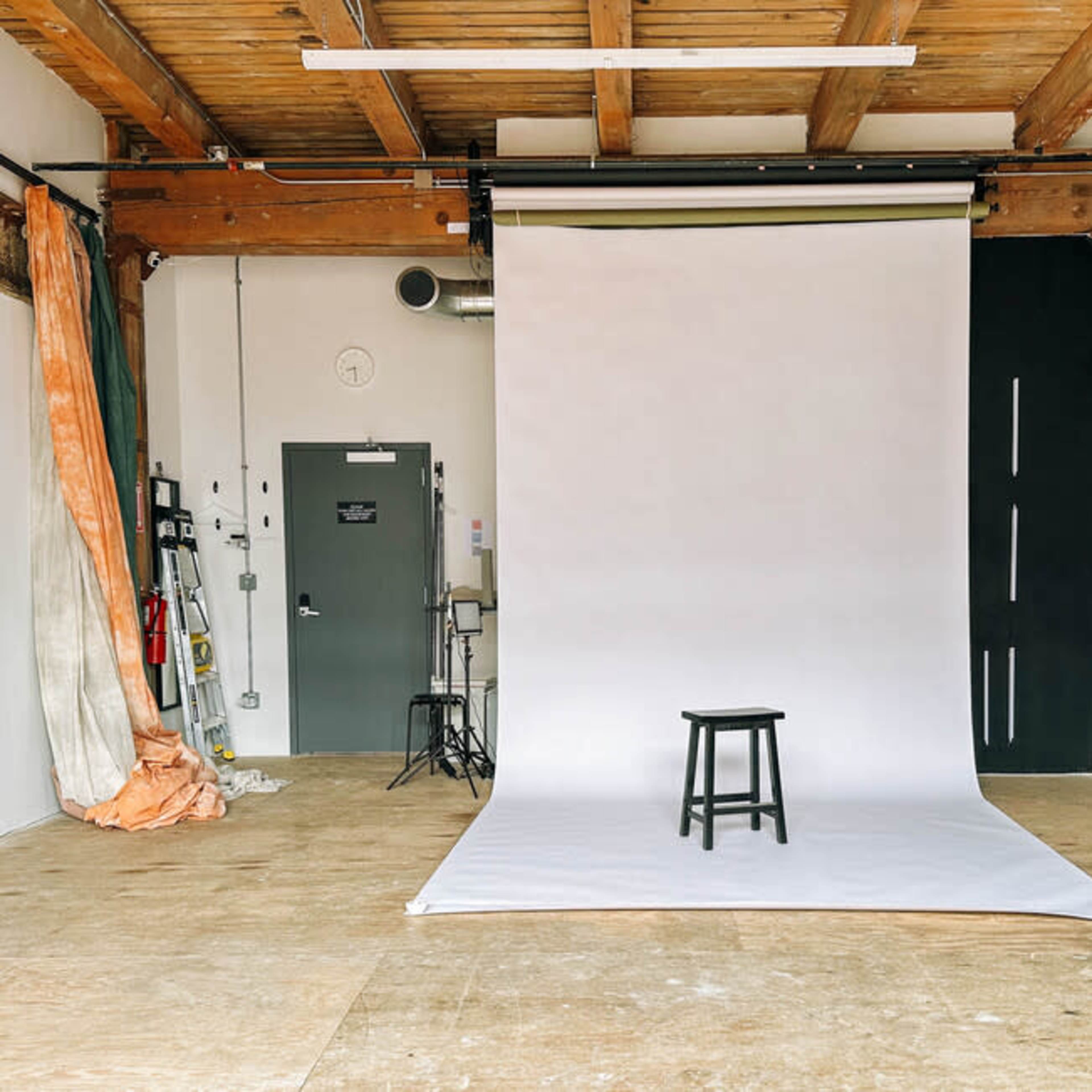 A minimalist photo studio with a wooden stool placed on a white backdrop and industrial-style walls.