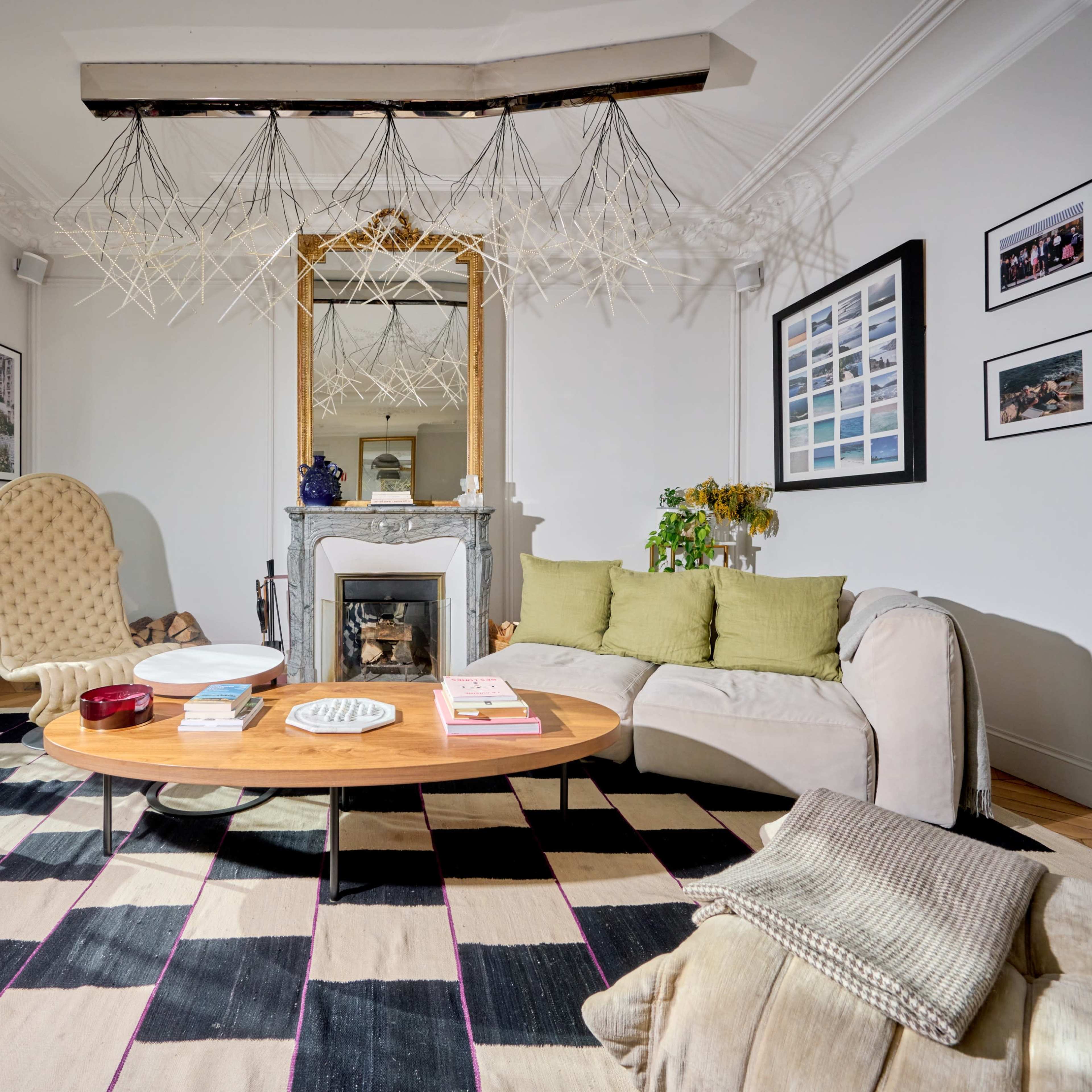 The living room features a round wooden coffee table, a beige sofa with green cushions, and a patterned black and white rug, with large windows and decorative wall art.