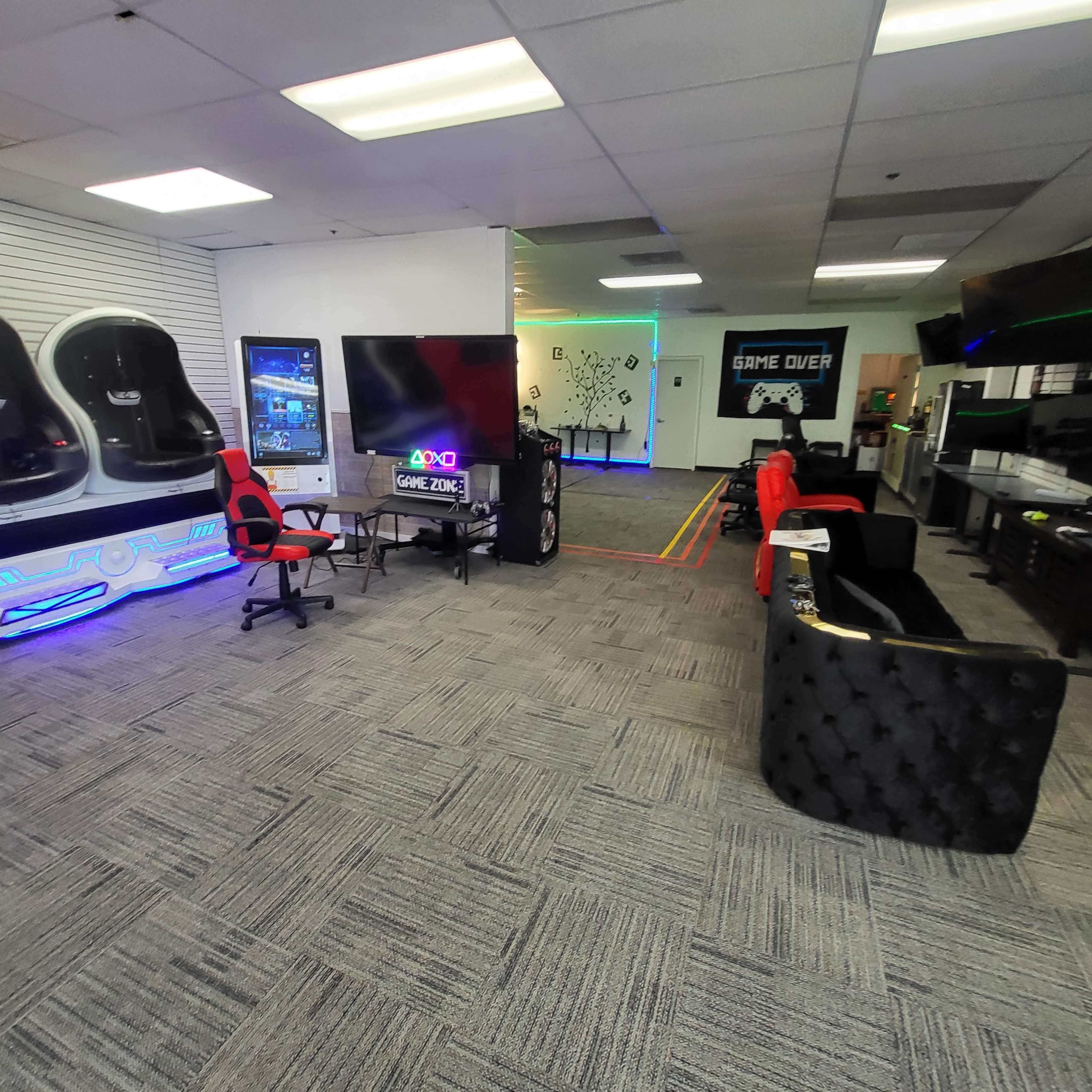 A gaming lounge with black and red furniture, a large TV, gaming machines, and a neon-lit wall featuring the phrase "GAME OVER."