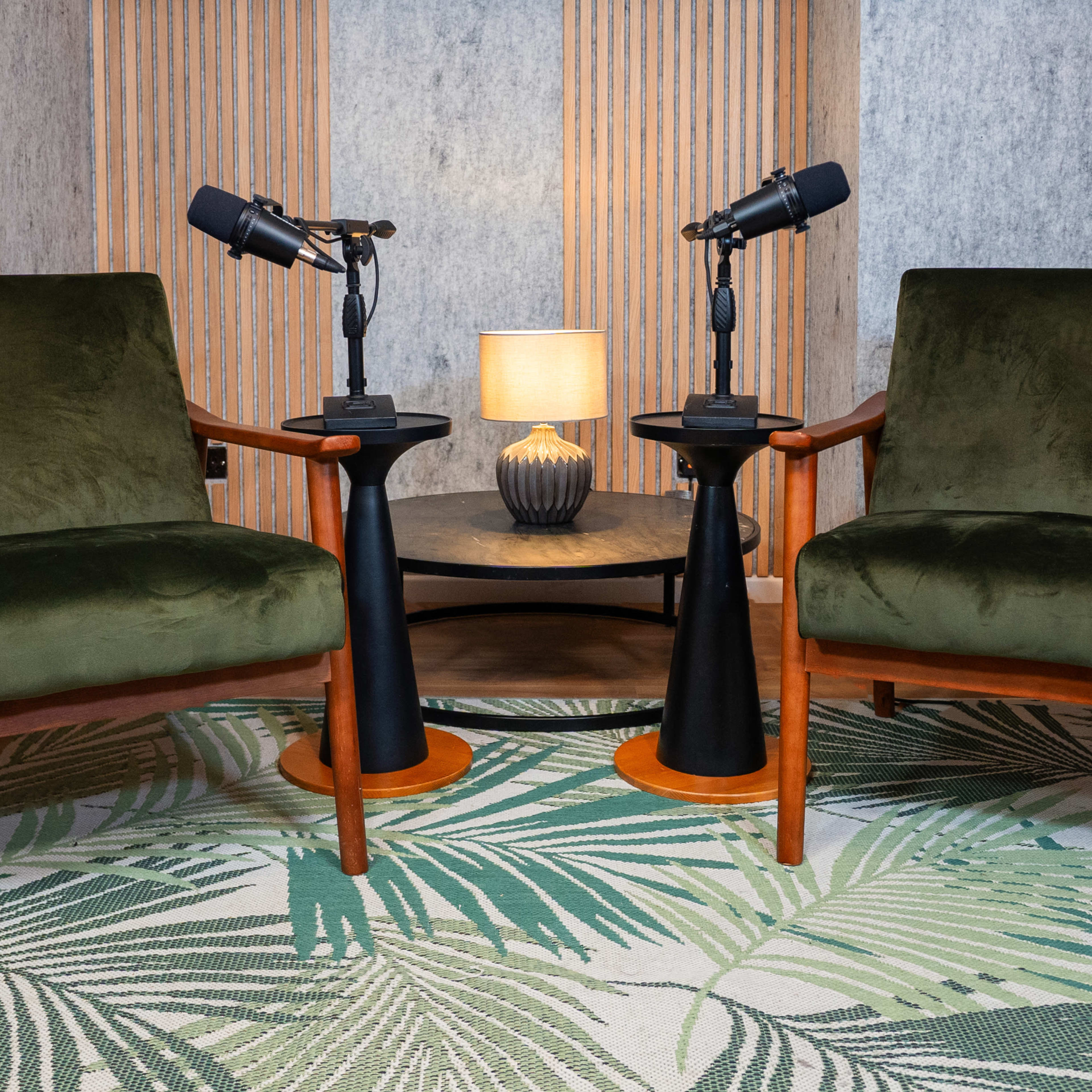 Two green upholstered chairs facing each other, positioned on a palm leaf-patterned rug, with a small table in between holding a lamp and two microphones.