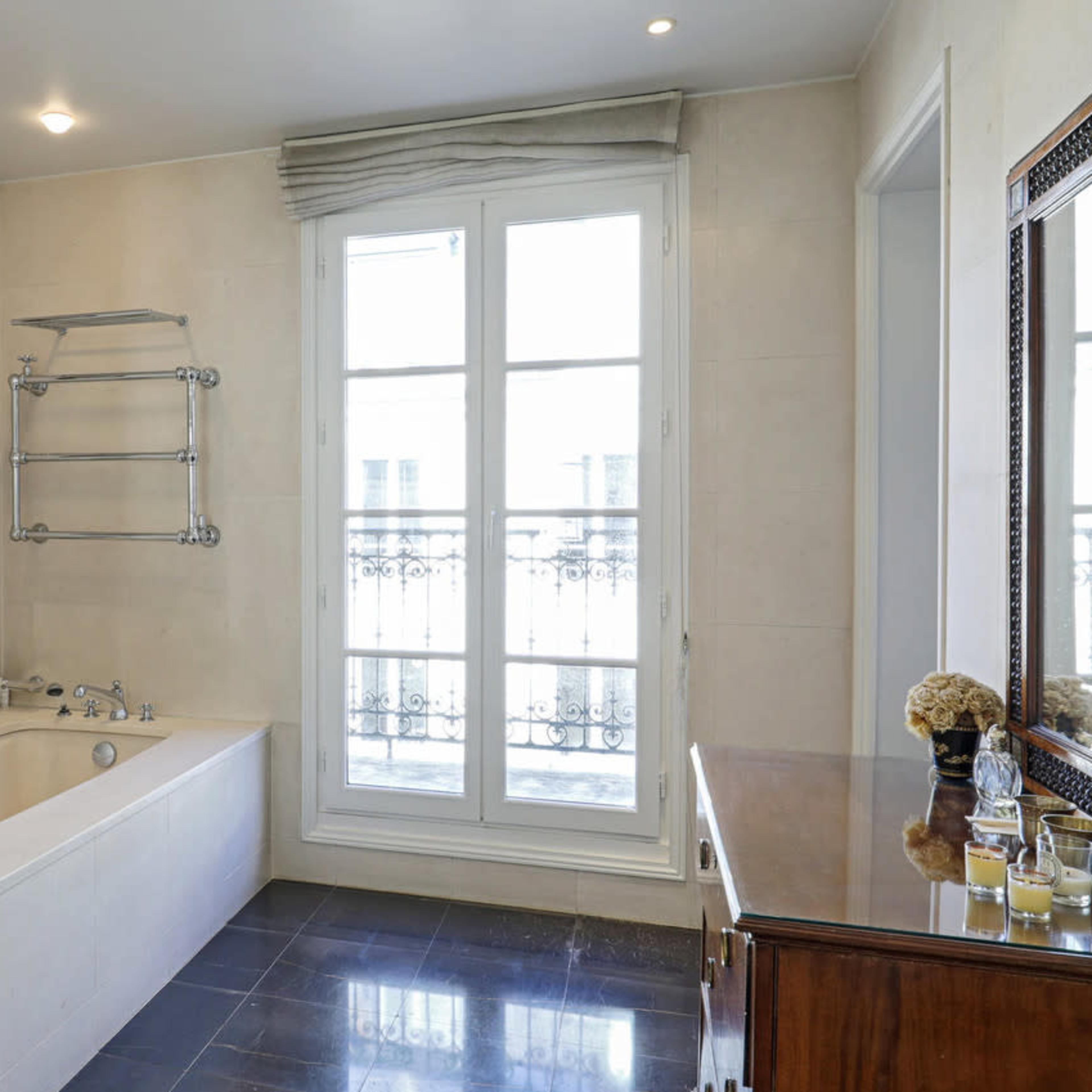 The bathroom features a freestanding bathtub, a wall-mounted towel rack, and a wooden vanity, with large windows providing natural light.