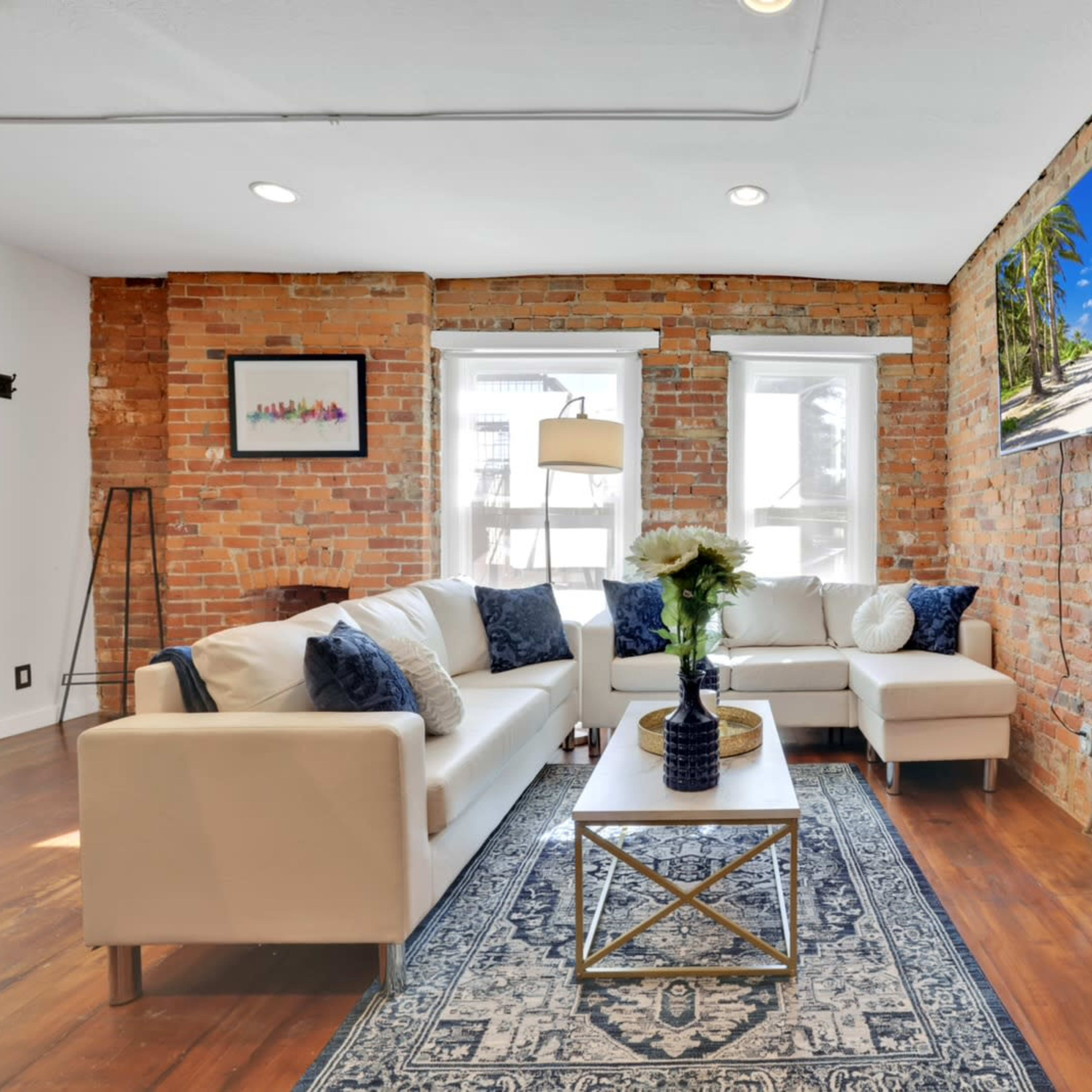 A living room with exposed brick walls, a white sofa, a coffee table, and a television mounted on the wall, featuring large windows that allow natural light.