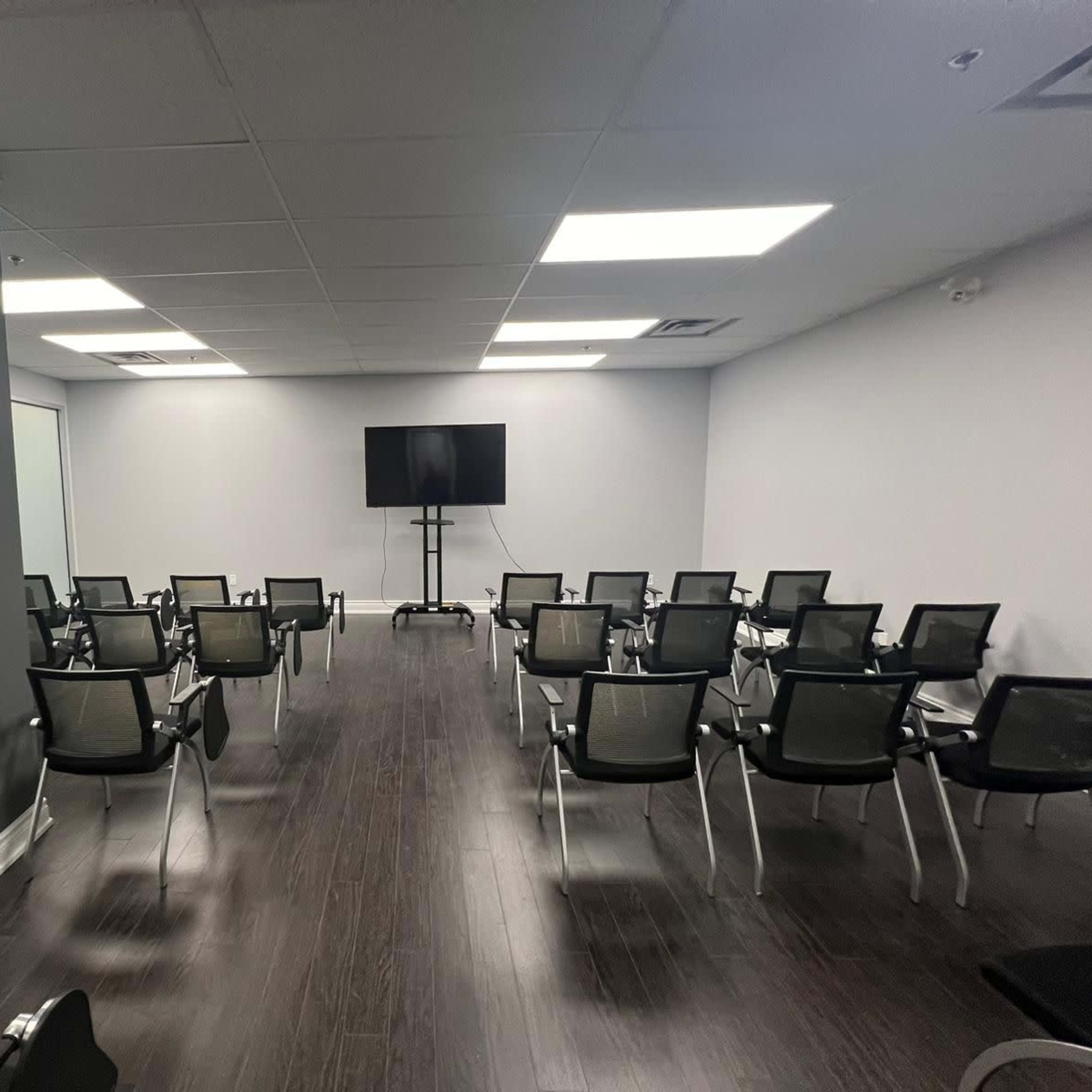 A conference room arranged with rows of black chairs facing a large wall-mounted screen.