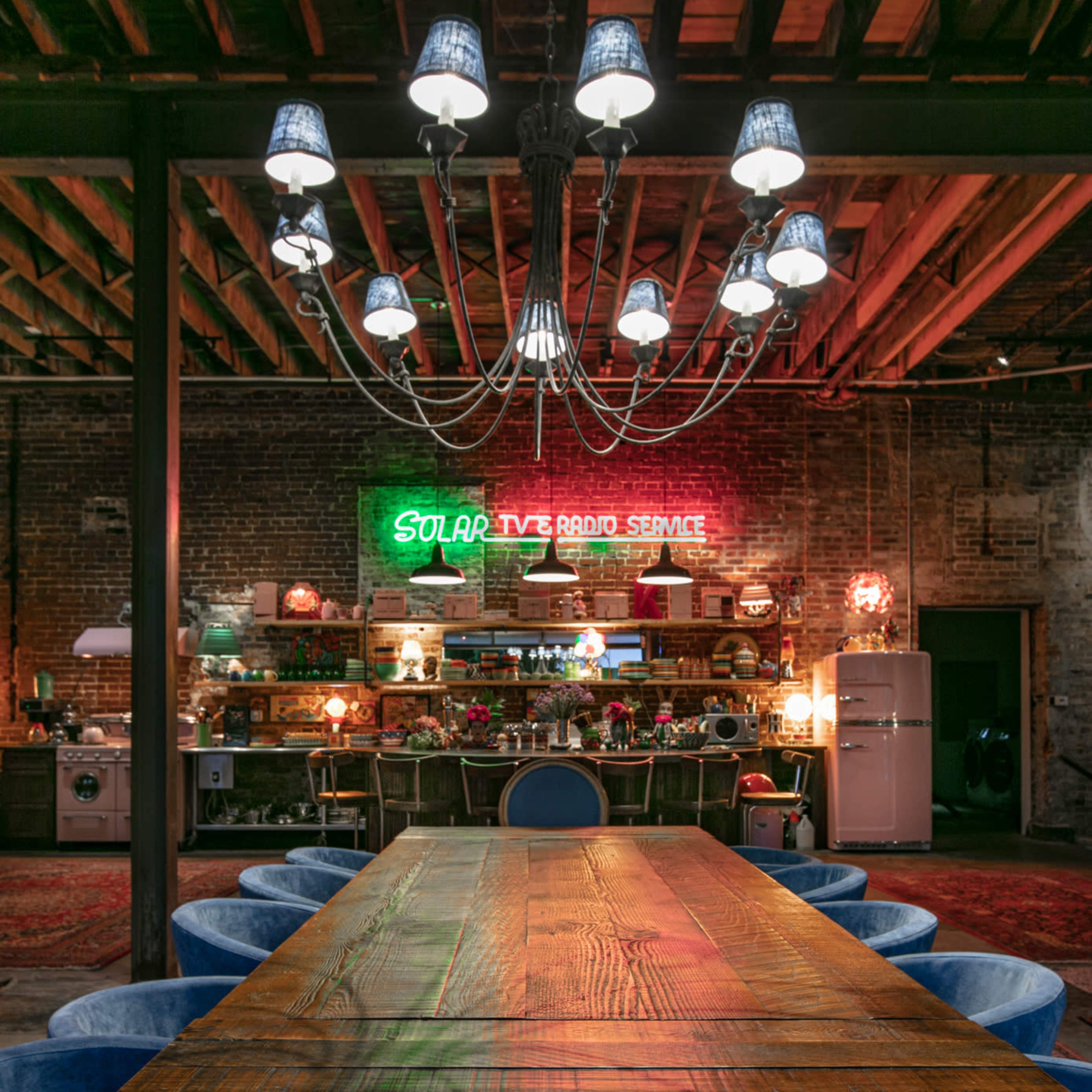A large wooden dining table is set in a rustic room with exposed beams and a neon sign that reads "Solar TV Radio Service" in the background.