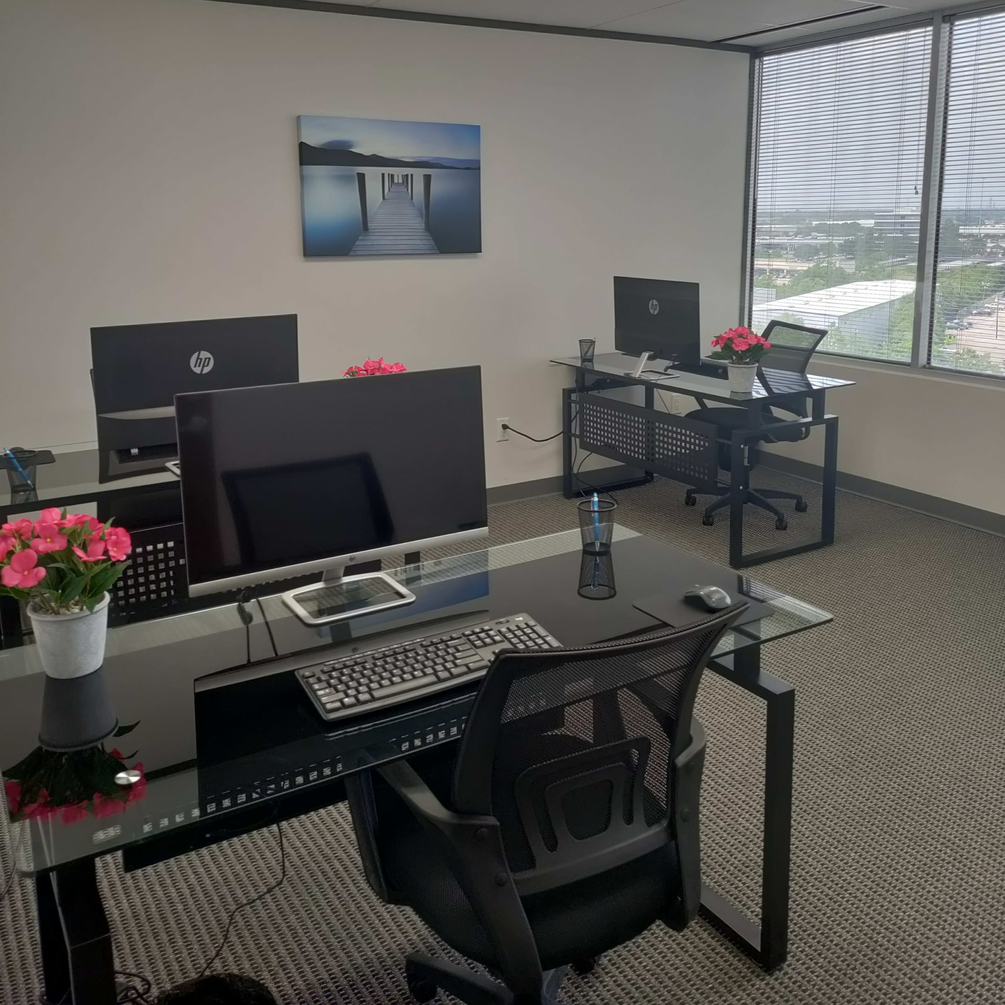 An office space with four workstations featuring computer monitors, keyboards, and potted plants, along with large windows offering a view of the outside.