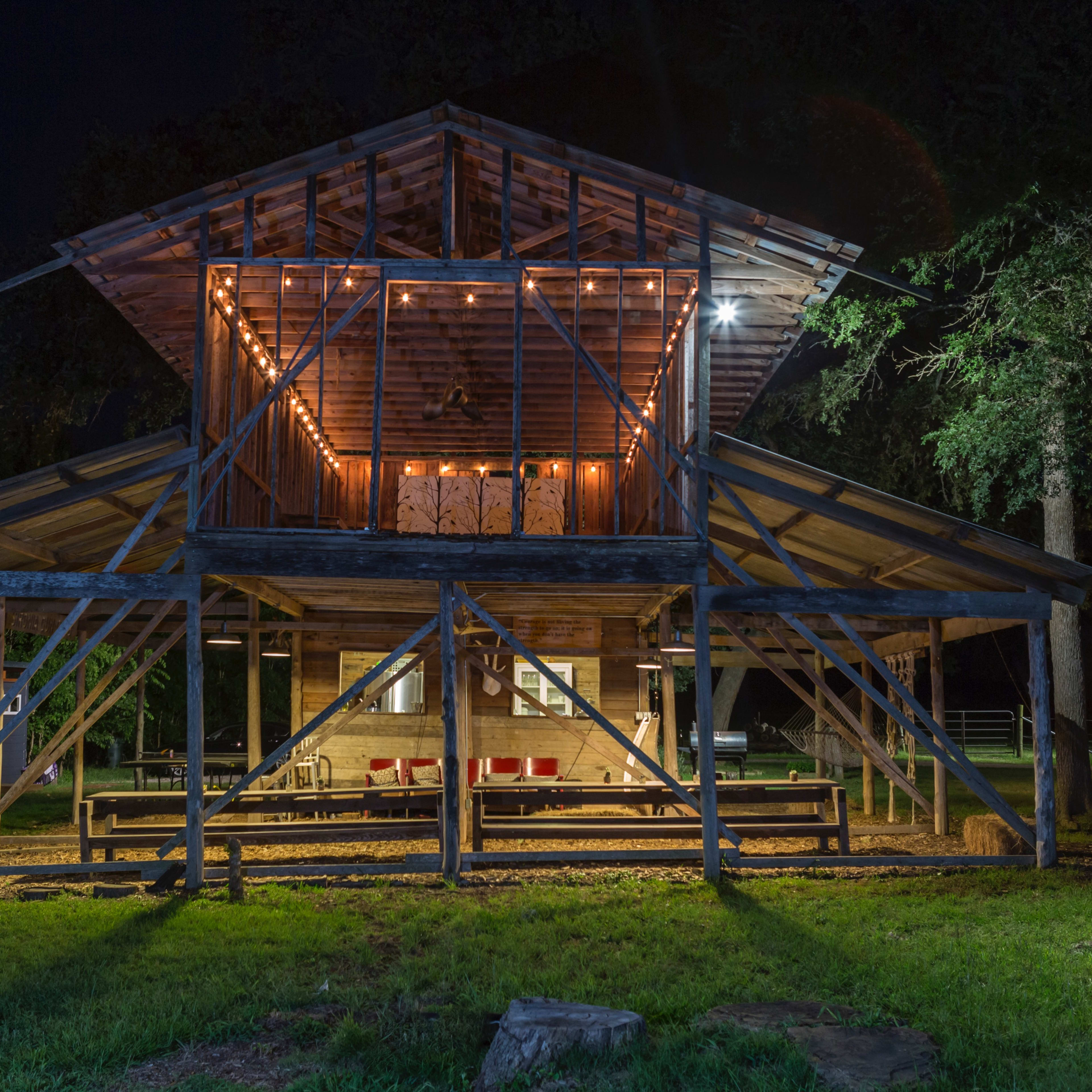 A wooden structure with an open lower level and a lofted upper area, illuminated by string lights at night, stands in a grassy environment.