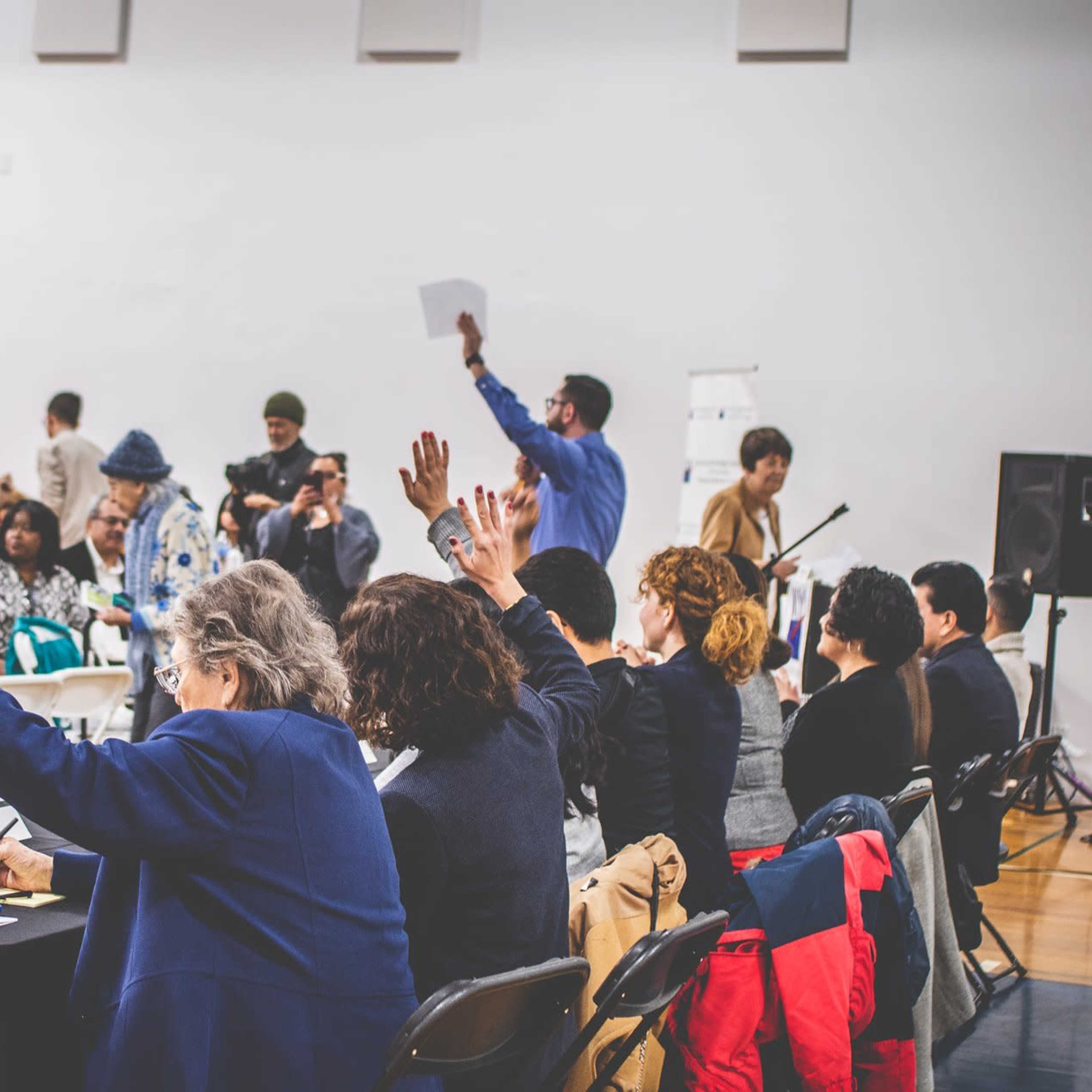 A group of people raises their hands in a large meeting space, while someone stands at the front holding a sheet of paper.