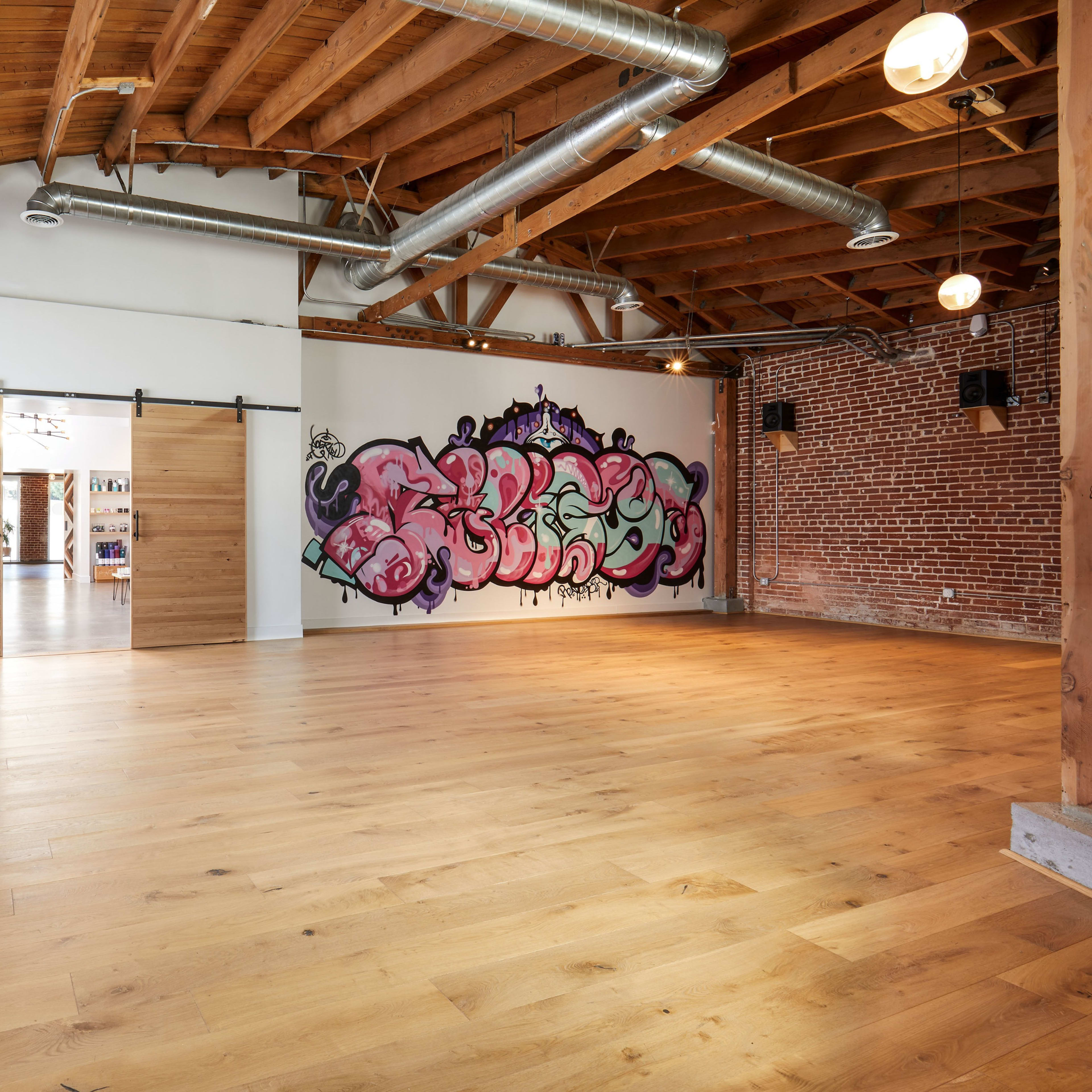 A spacious interior room with wooden floors, exposed brick walls, and a large mural on one wall, complemented by sliding wooden doors.