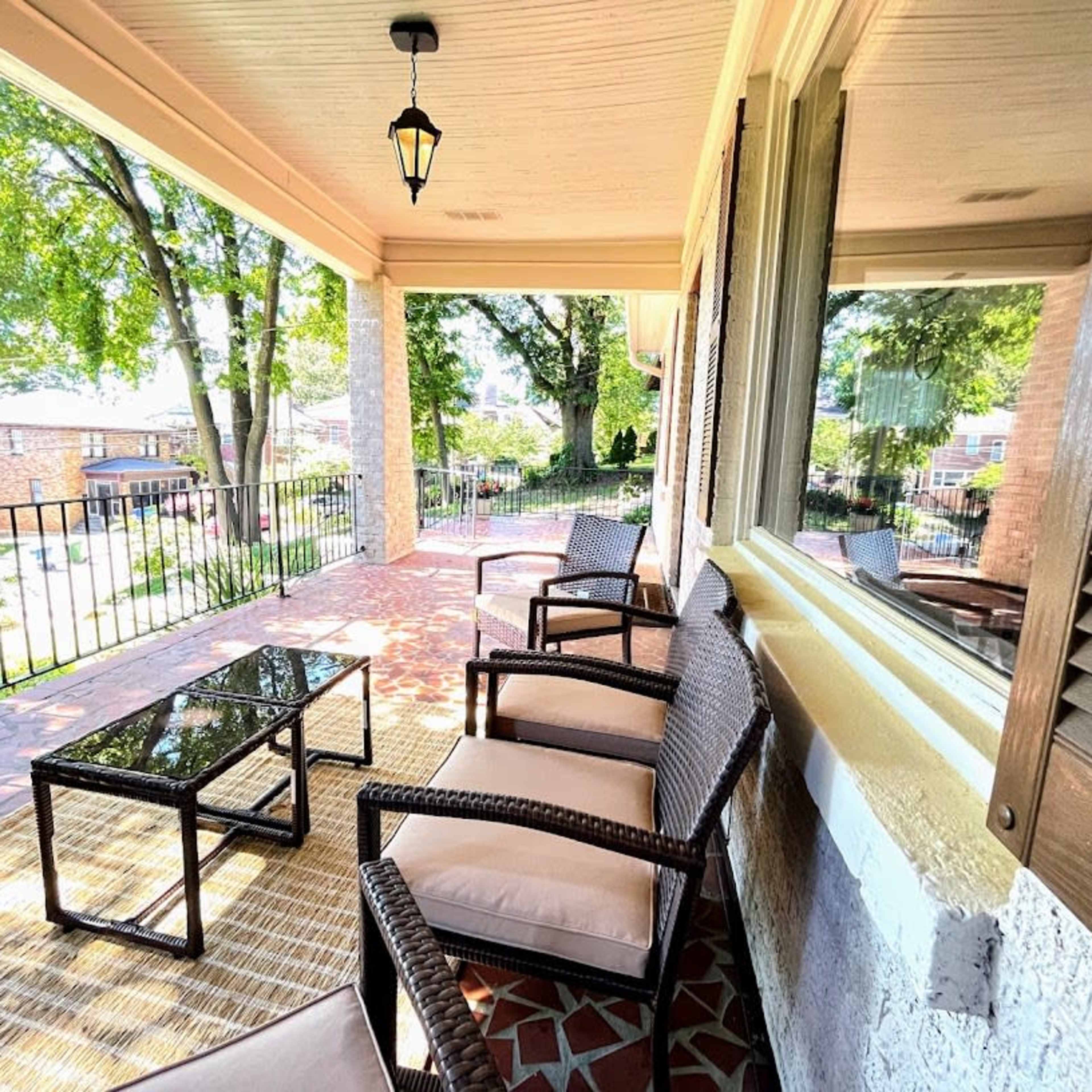 A covered porch with several chairs and a glass-top coffee table, surrounded by greenery and visible houses beyond a railing.