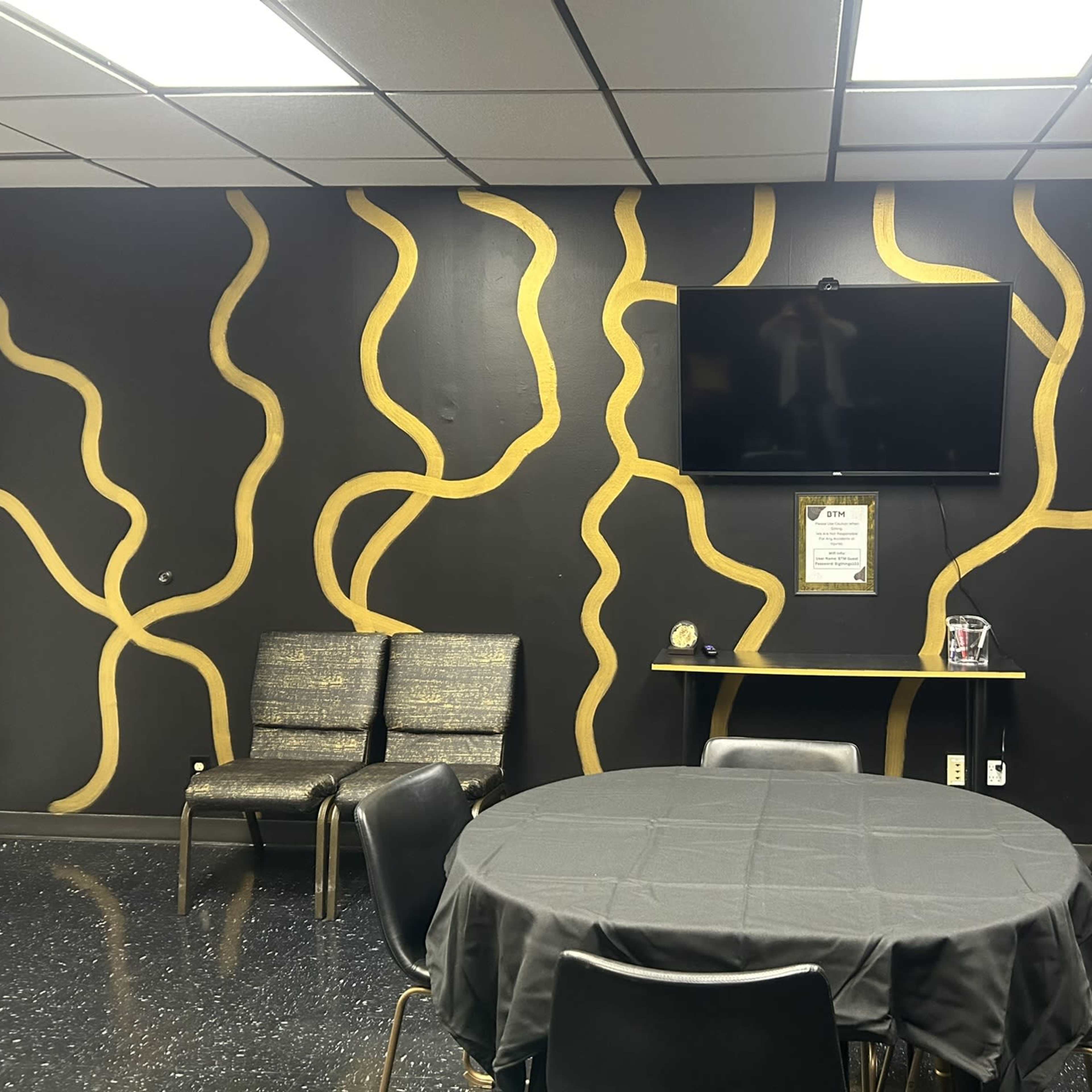 A room with a round table covered by a black tablecloth, a television mounted on a wall with a patterned design, and several chairs arranged nearby.