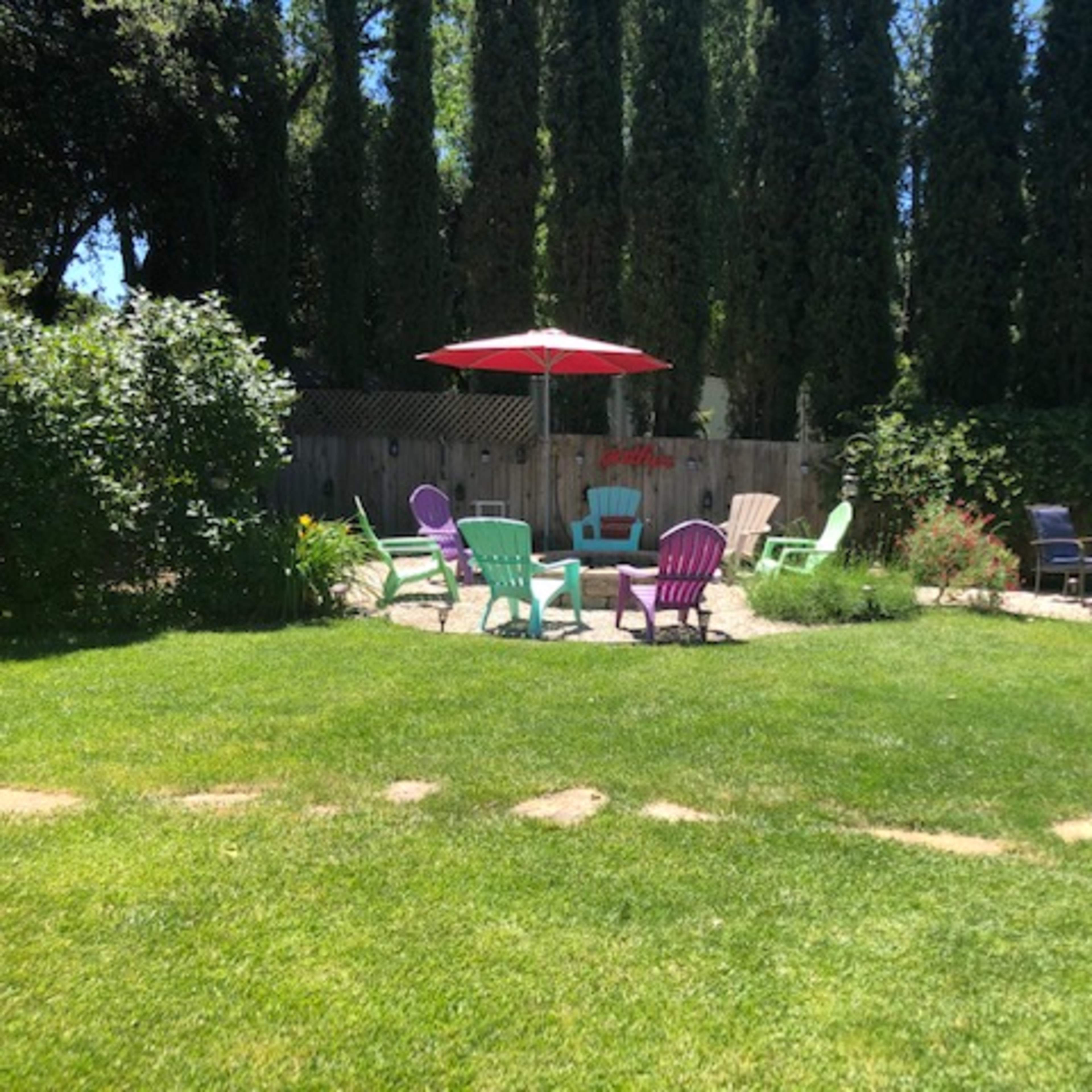 A landscaped yard with colorful chairs, a red umbrella, and neatly arranged gravel pathways surrounded by tall trees and greenery.