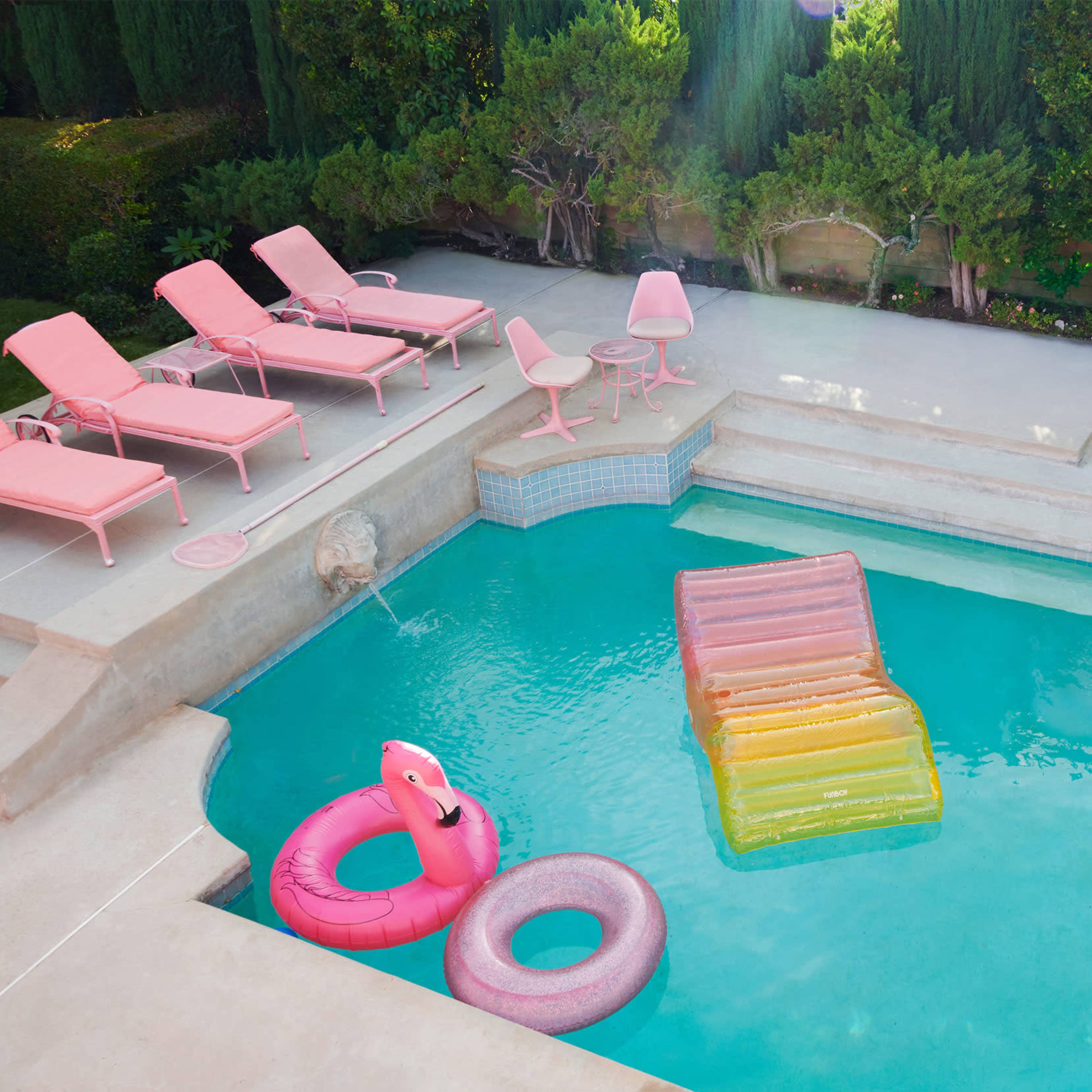 A swimming pool with a colorful inflatable slide, a pink flamingo float, and pink lounge chairs arranged on a deck surrounded by greenery.