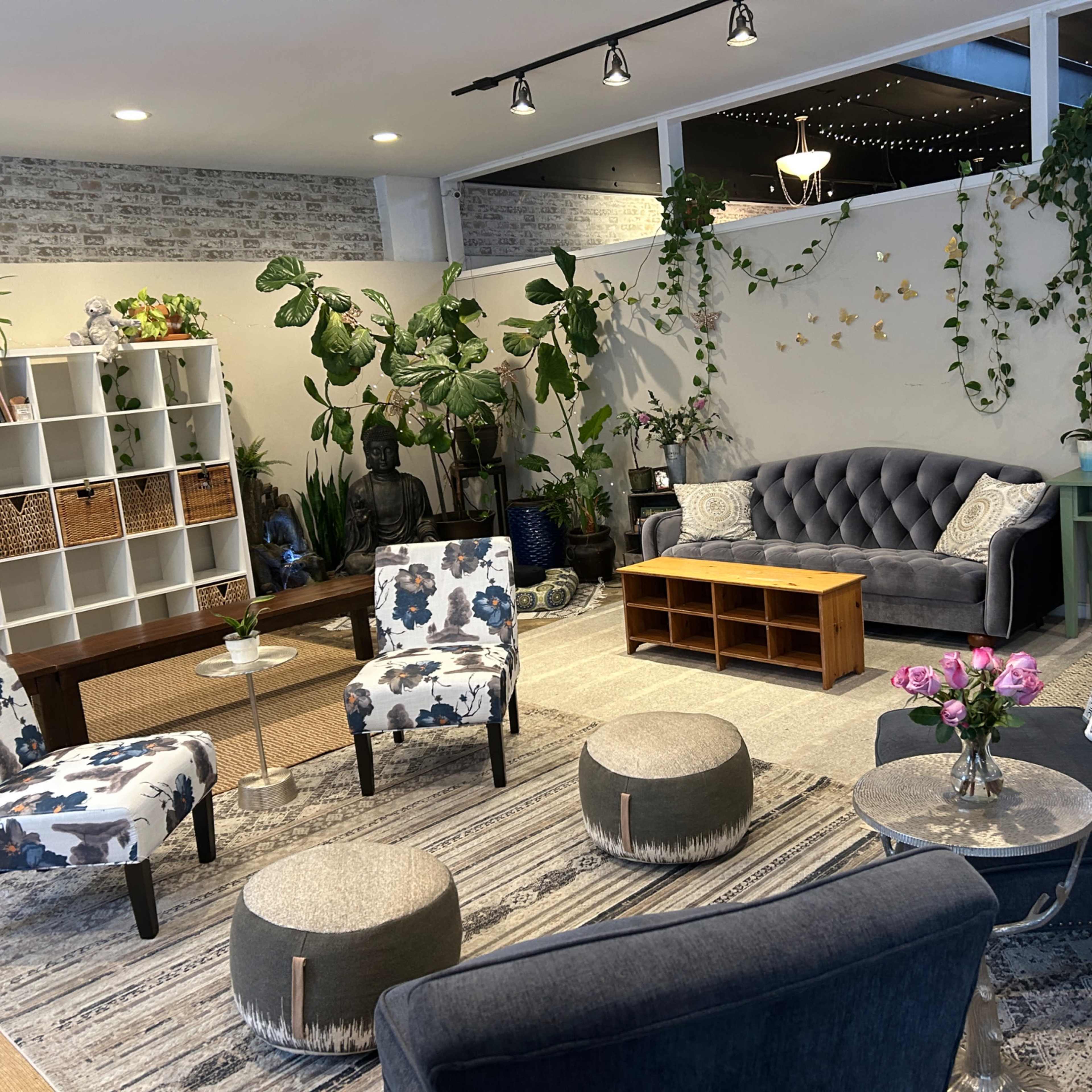 A cozy lounge area with various seating options, green plants, and decorative shelves against a light-colored wall.