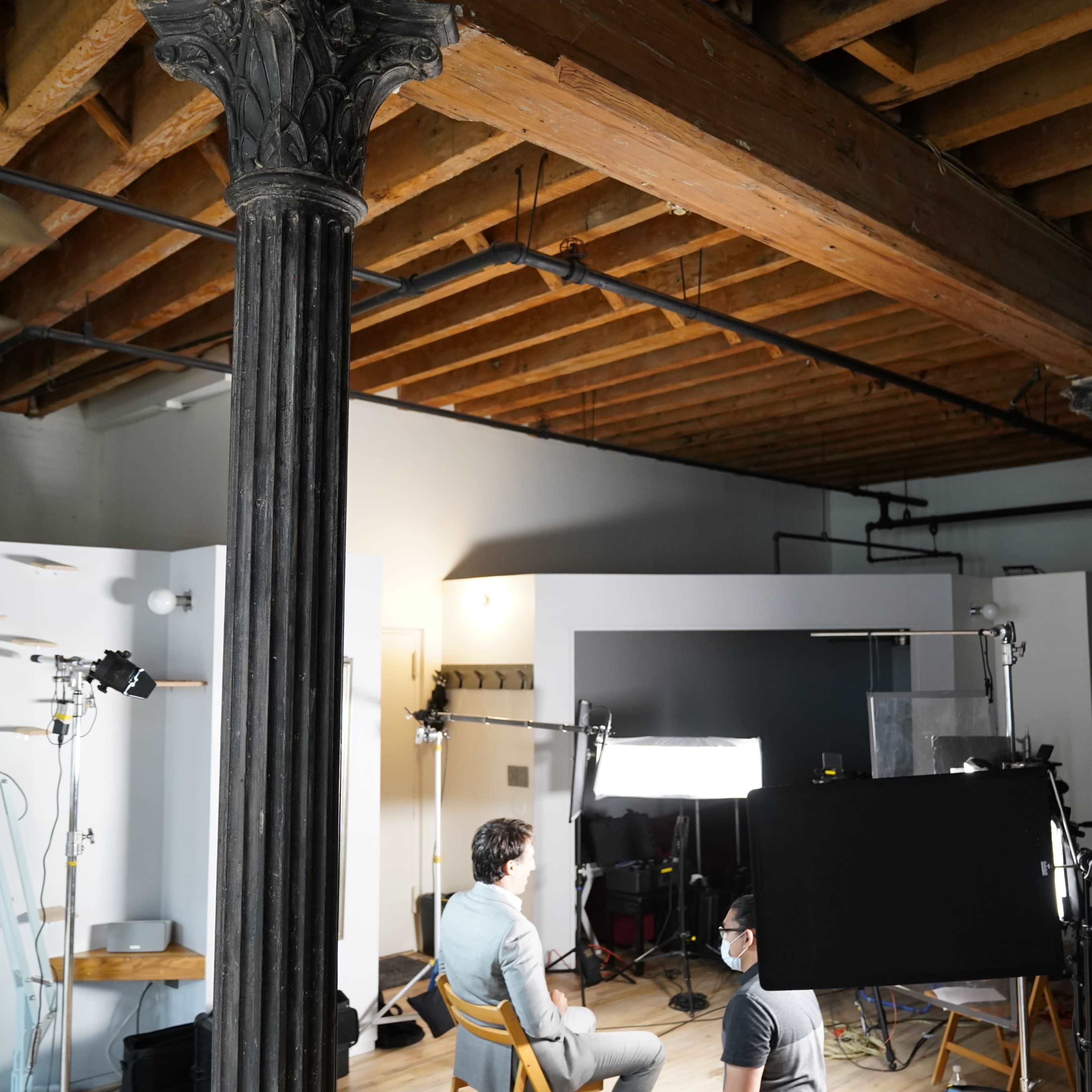 A film crew sets up equipment in a spacious studio with wooden beams and columns while two people sit in front of a black backdrop.