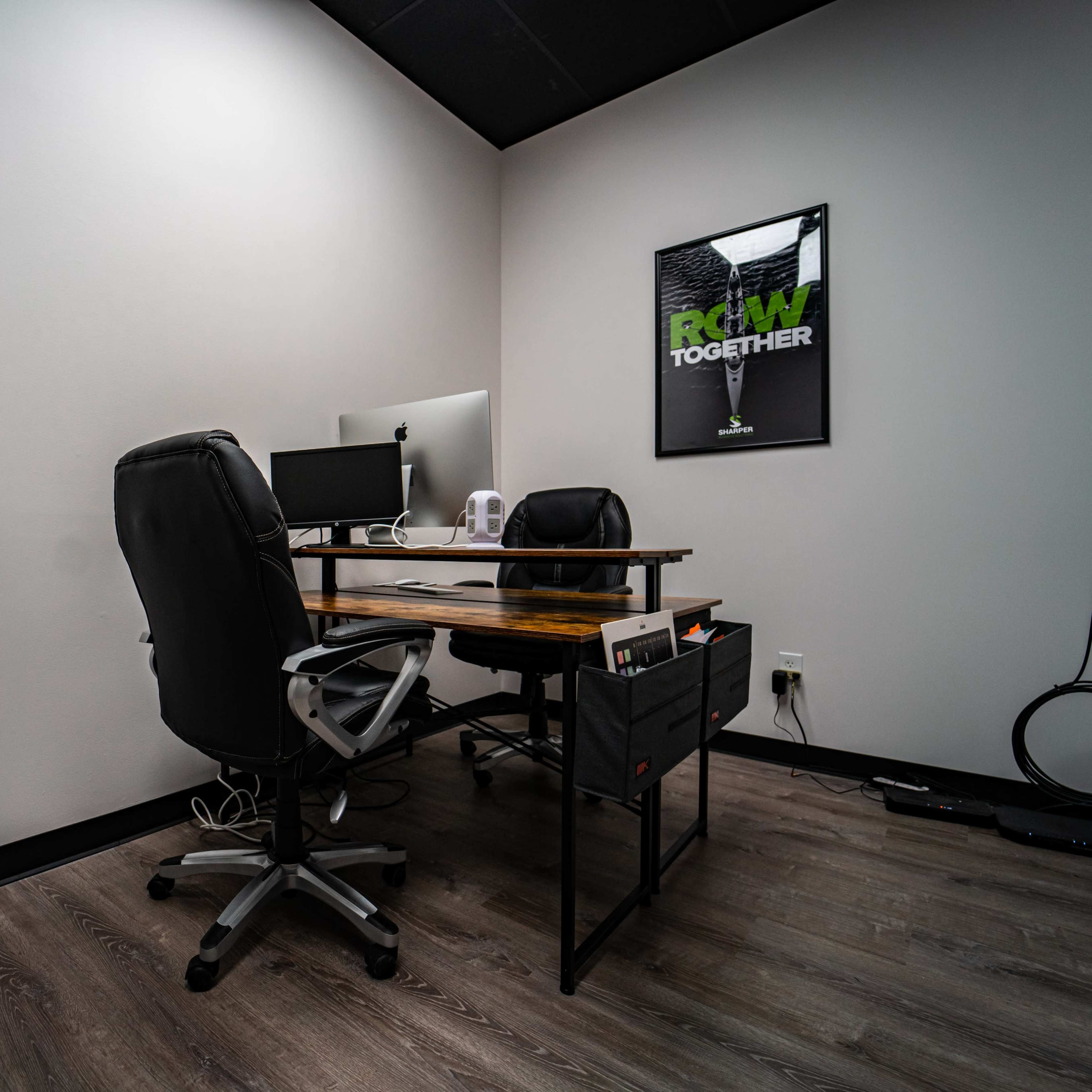 A minimalist office space featuring a wooden desk with two office chairs, a computer, and a motivational poster on the wall.