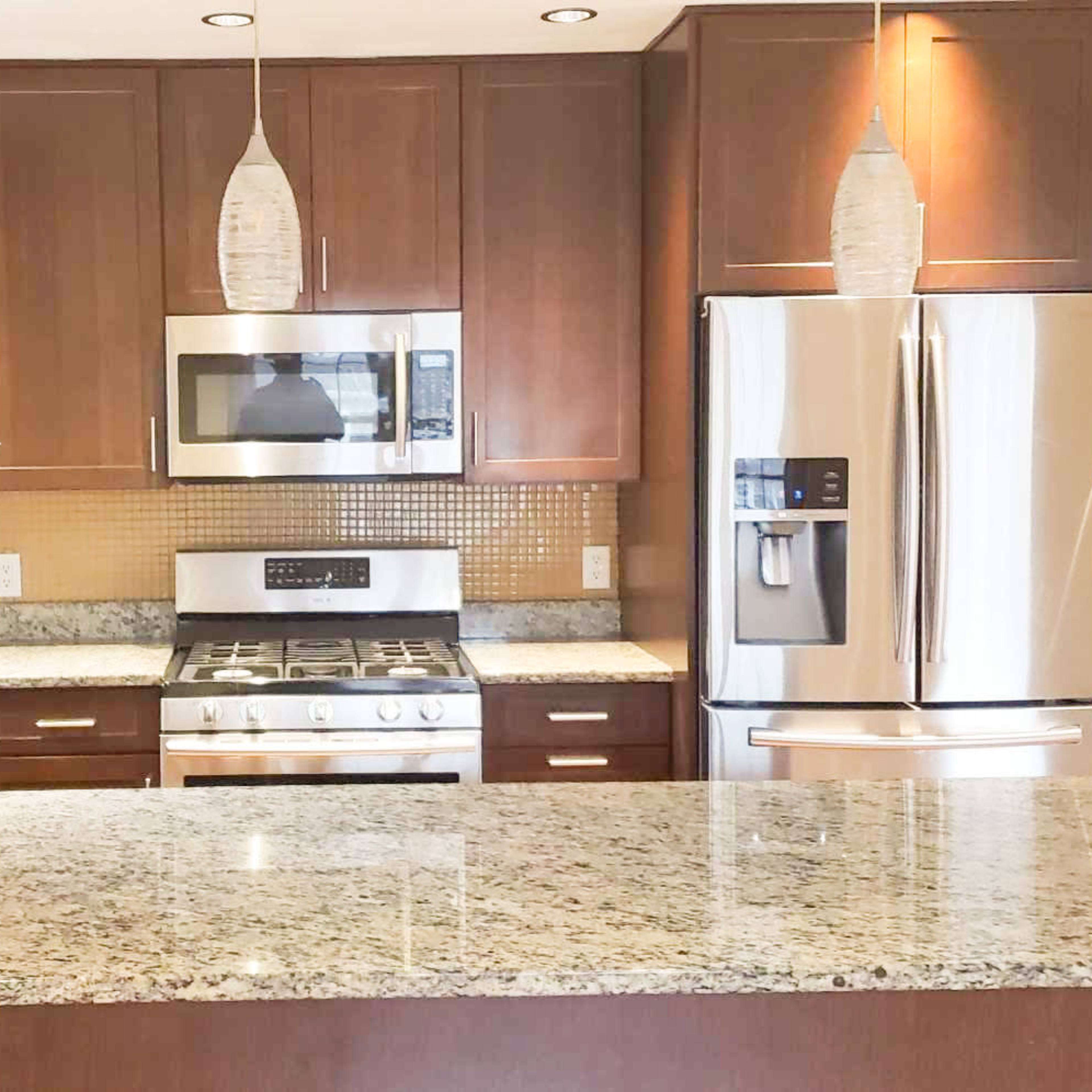 A modern kitchen featuring brown cabinetry, stainless steel appliances, a granite countertop, and pendant lighting.