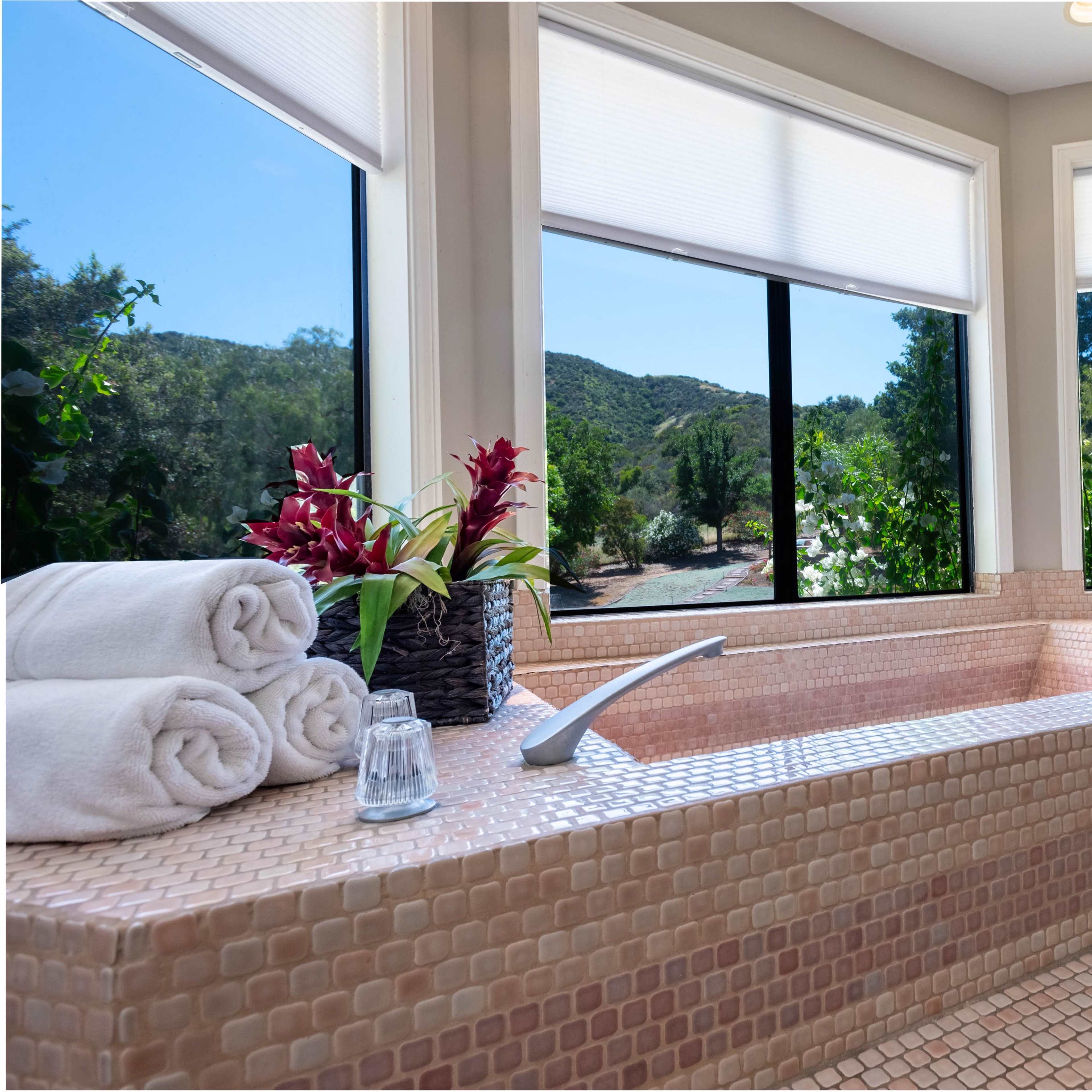 A modern bathroom features a large tub with neatly stacked towels and a scenic view of greenery through the windows.