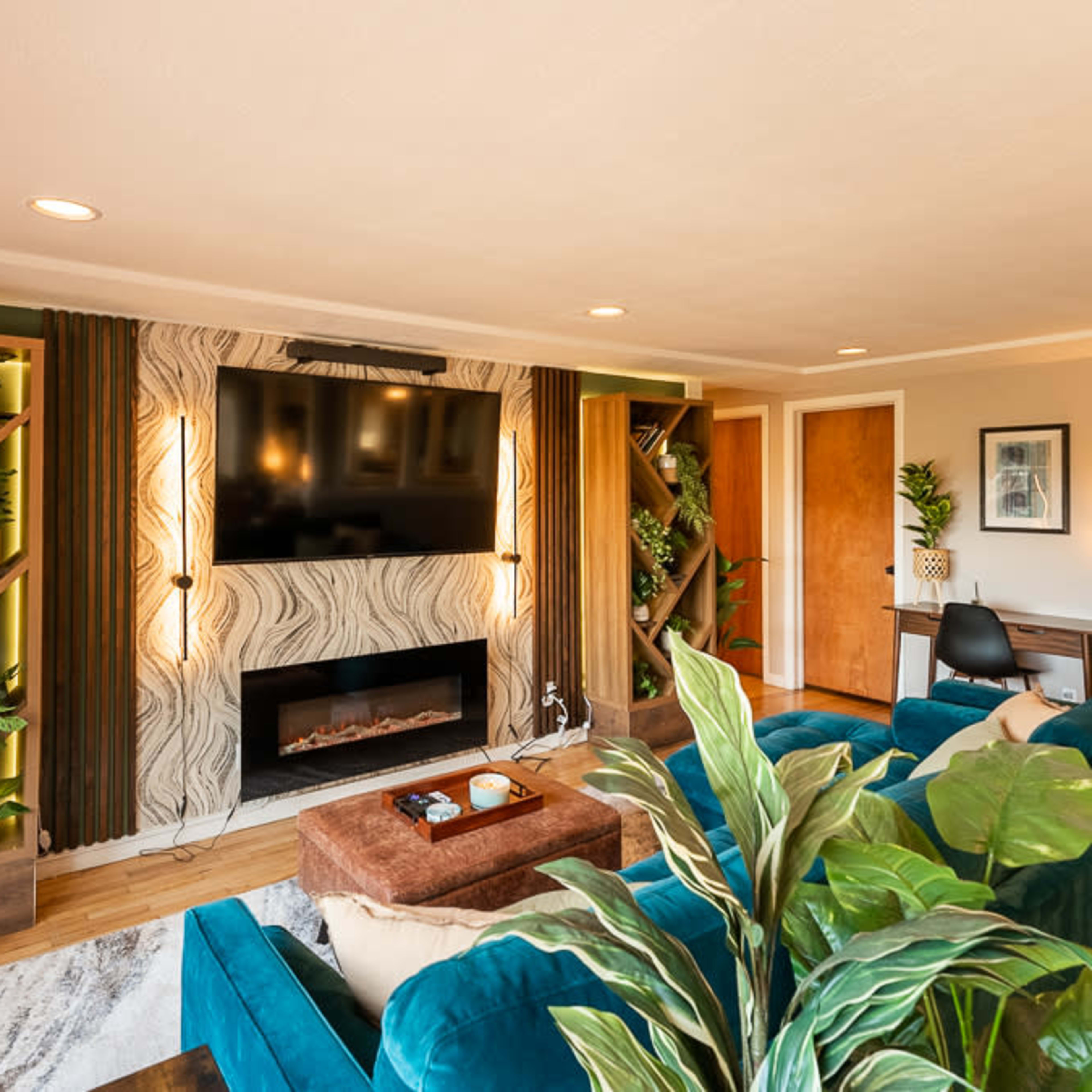 A modern living room featuring a large TV mounted on the wall, surrounded by green plants and built-in shelving, with a couch and a decorative coffee table in the center.