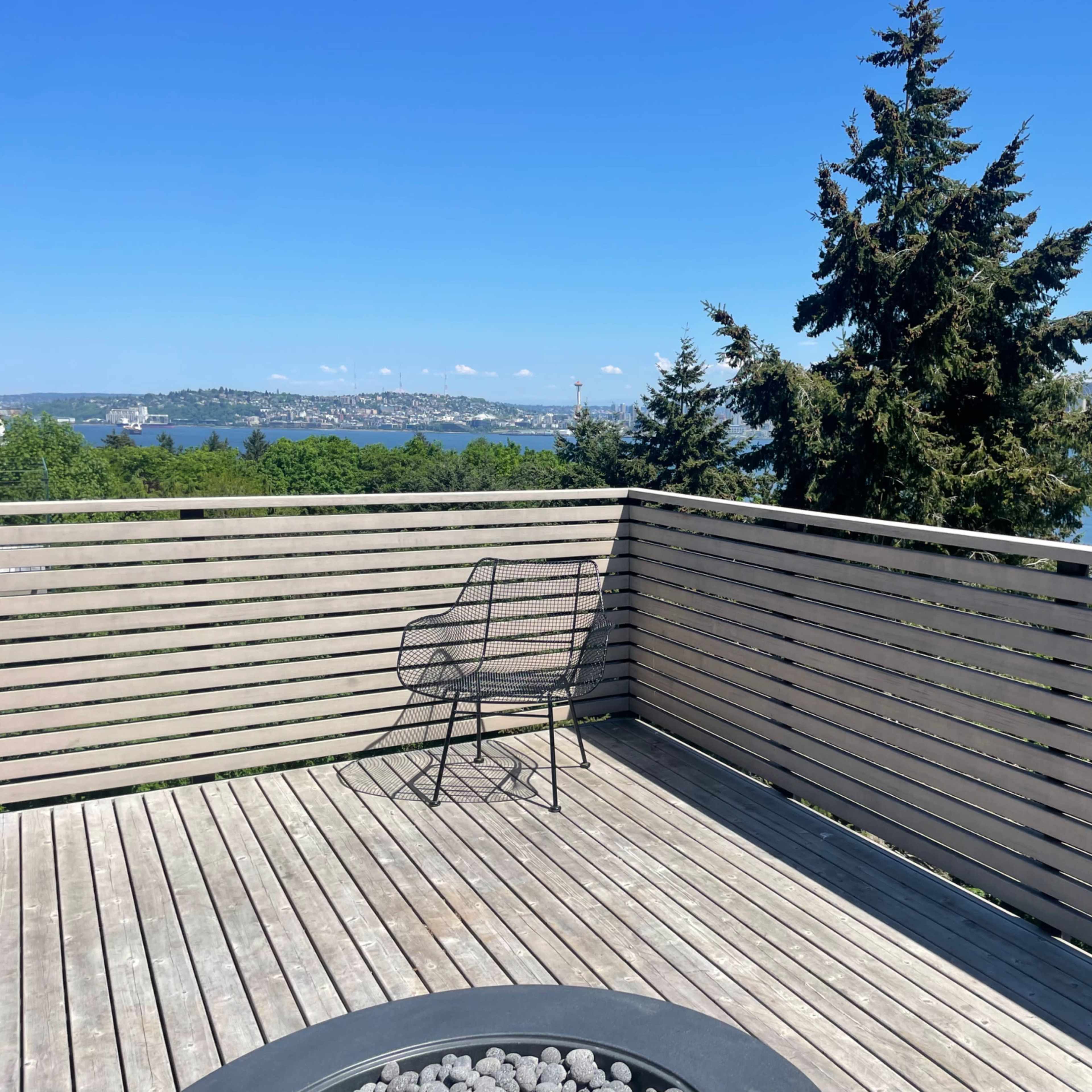 A wooden deck with a metal chair and a circular fire pit overlooks a body of water and trees, under a clear blue sky.