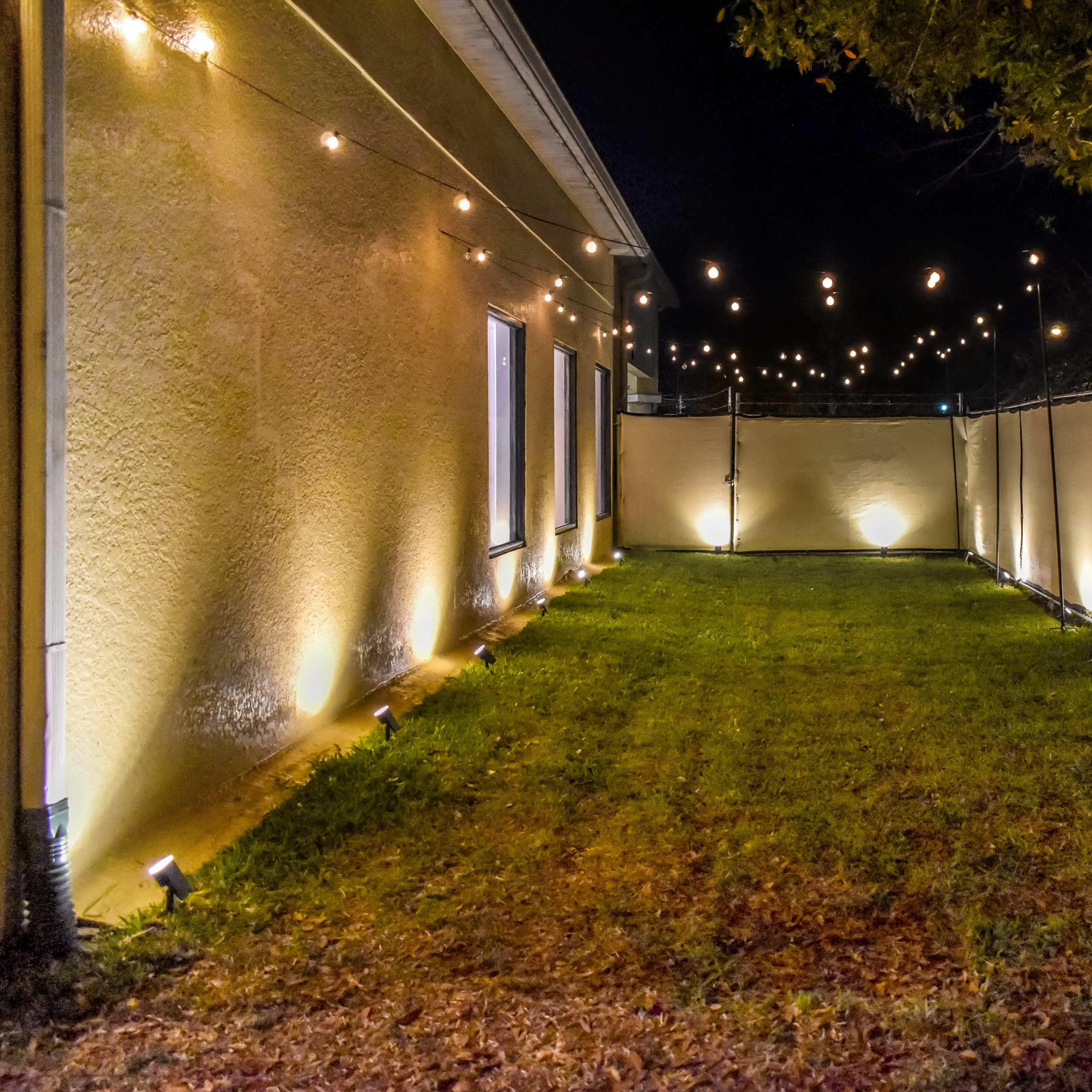 A yard illuminated by string lights and garden lights, enclosed by a fence and a building at night.
