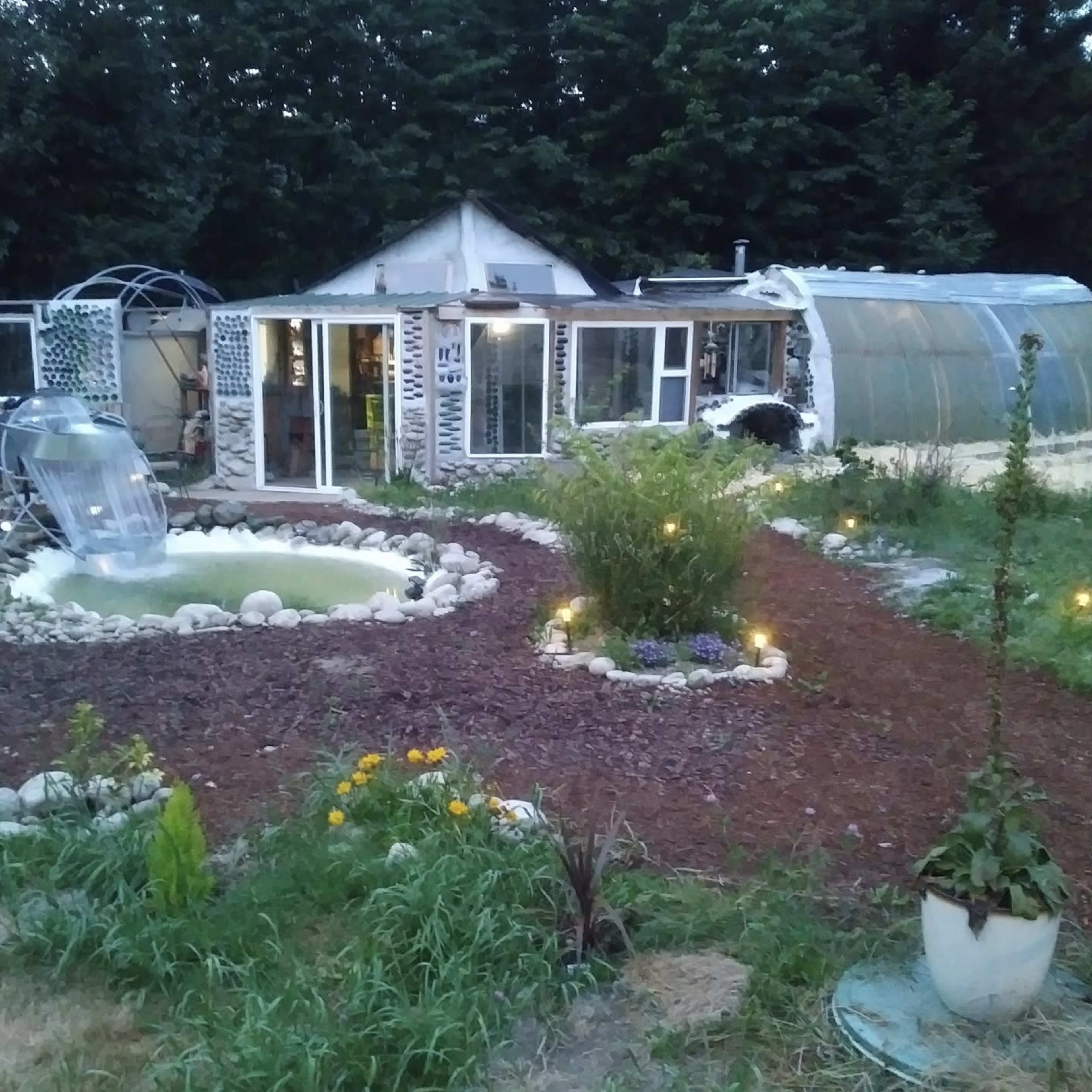 A garden with a small pond and a water slide, surrounded by a rocky path, leading to a greenhouse and a house in the background.