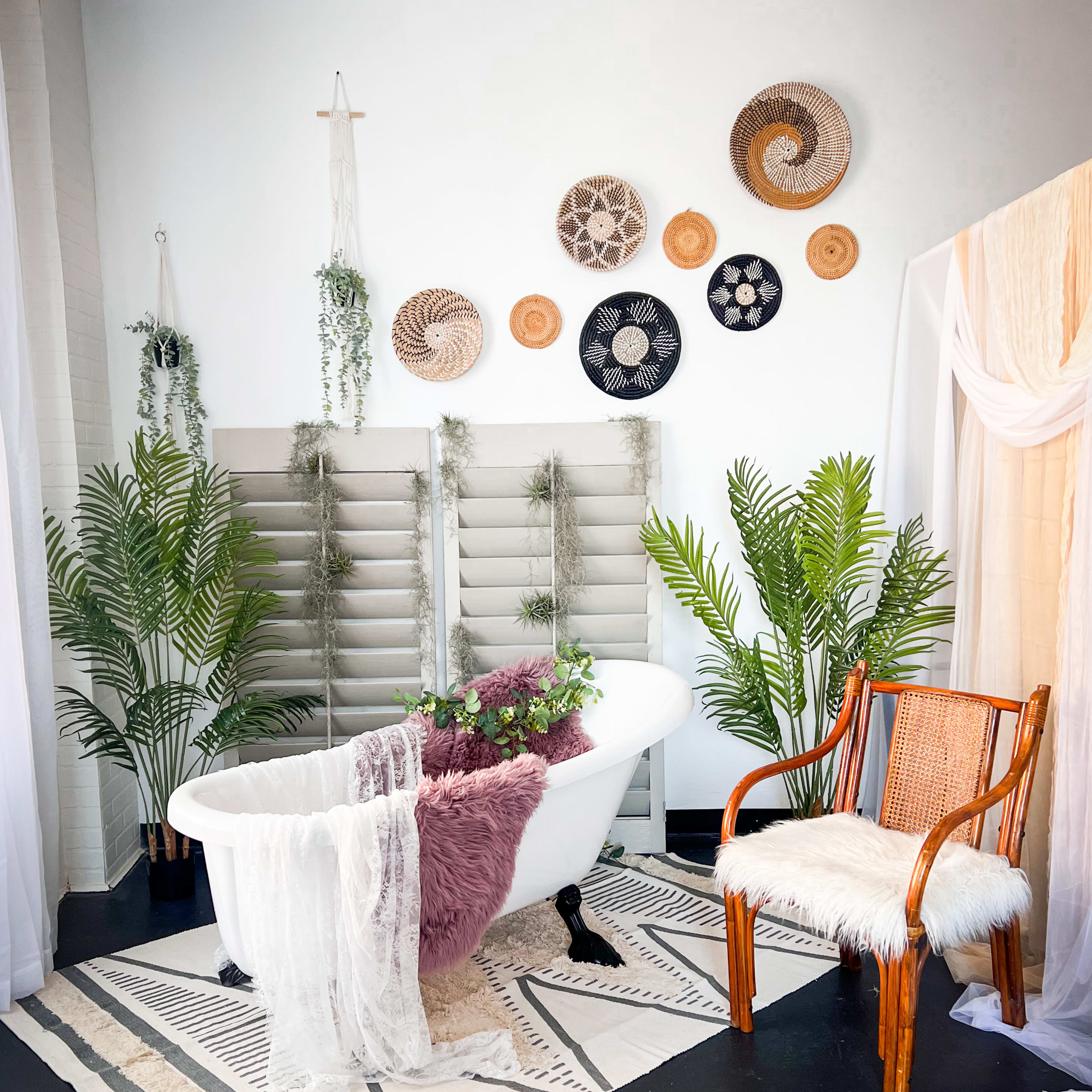 A stylish bathroom with a freestanding white bathtub, a rustic chair, decorative wall hangings, and greenery.