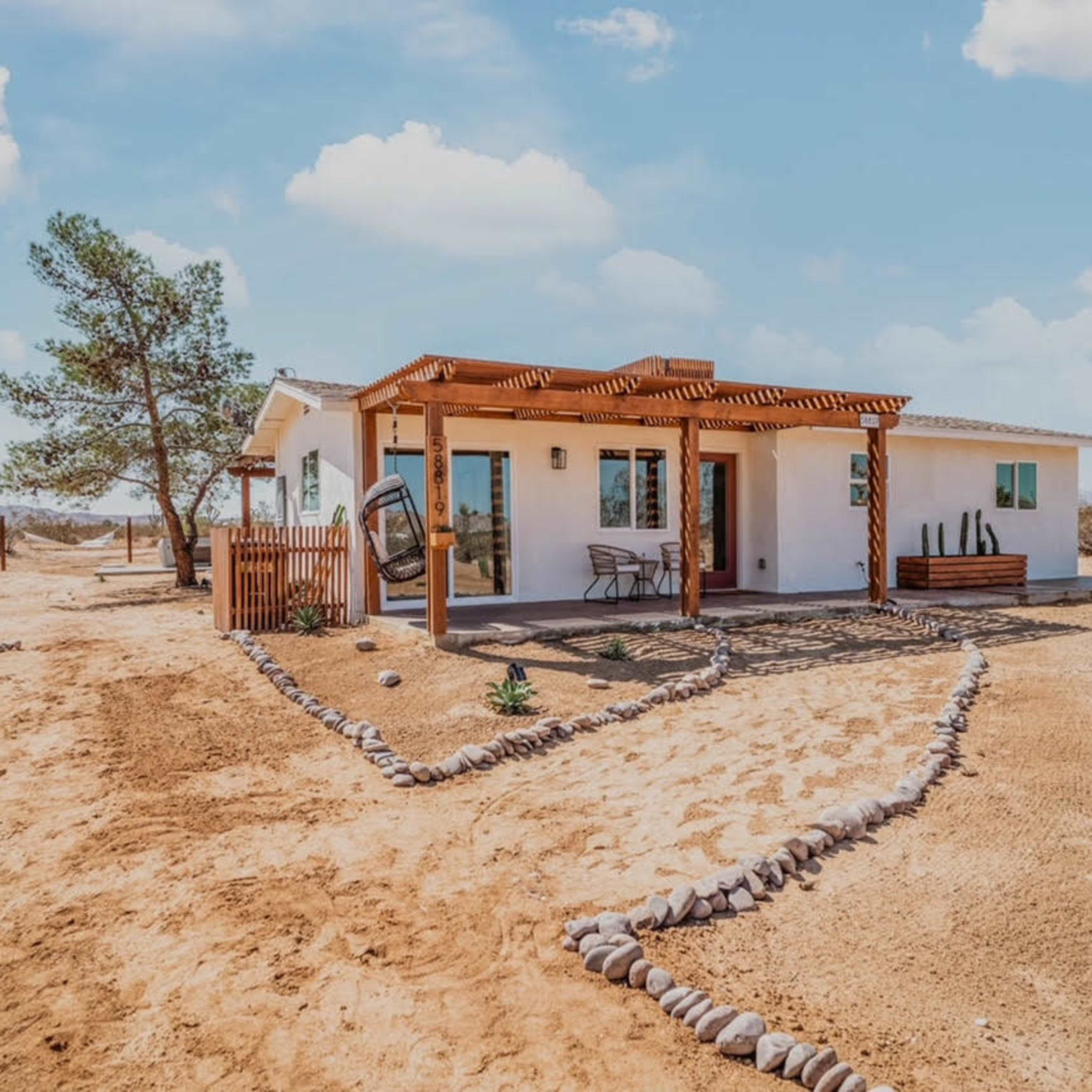 A small, modern house with a wooden pergola is set against a desert landscape with sparse vegetation and a clear blue sky.