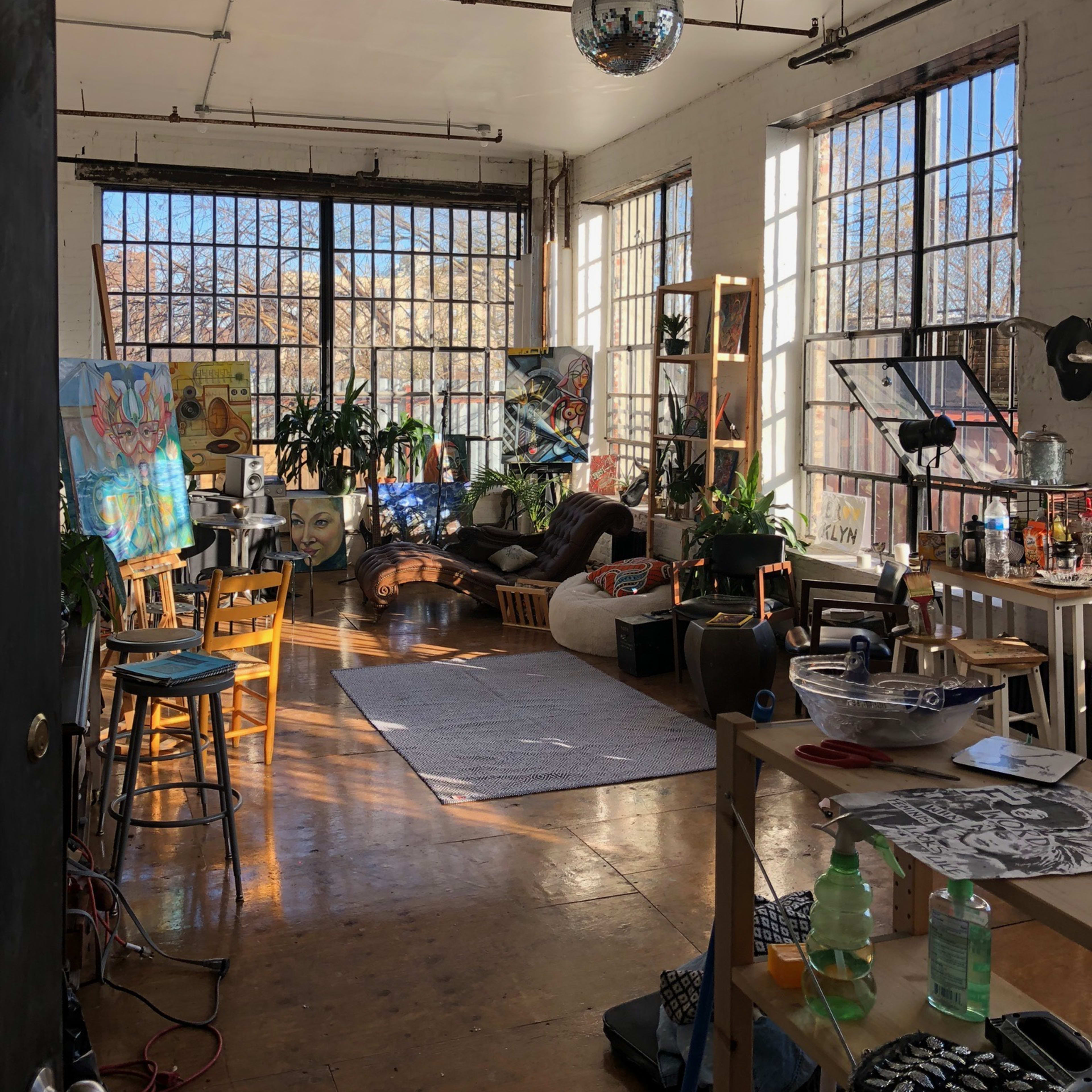 An open artist studio with large windows, various plants, artwork on the walls, and a mix of seating and workspaces.