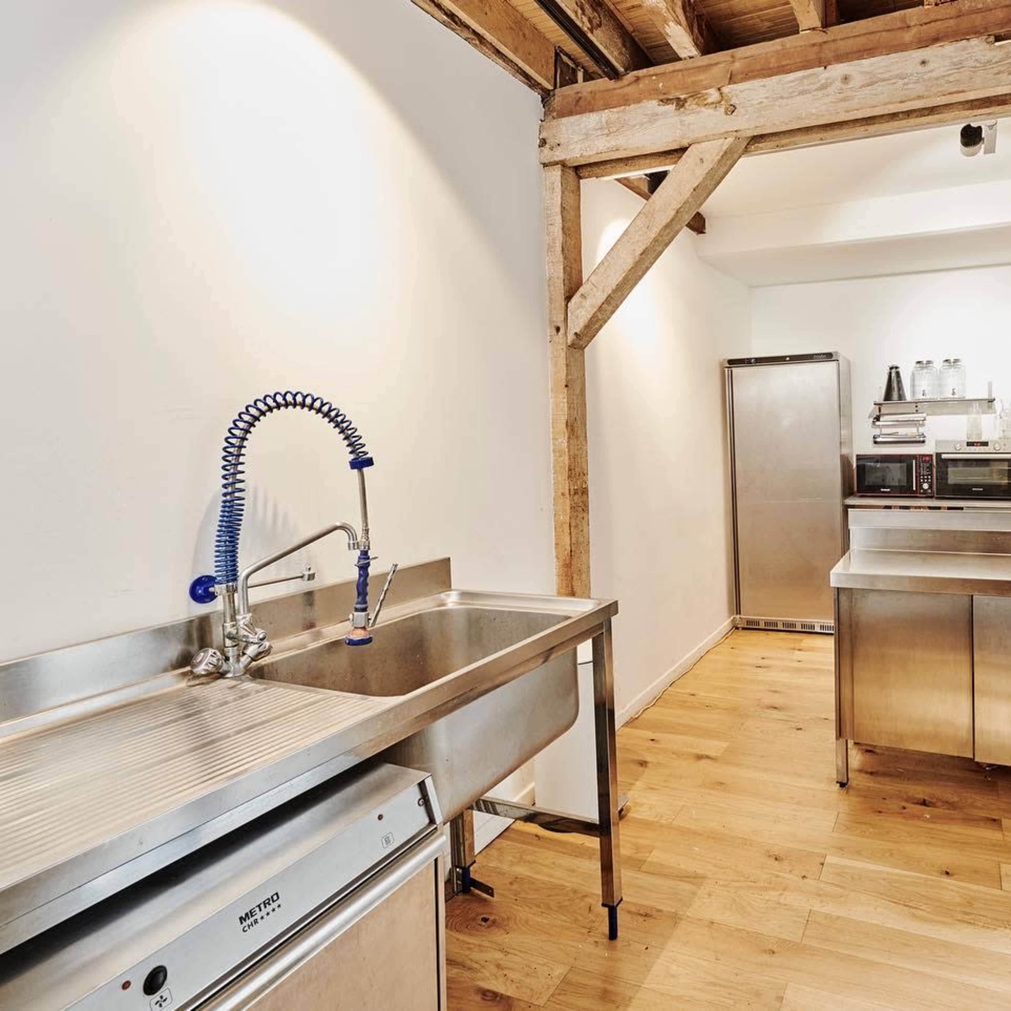 A modern kitchen with stainless steel appliances, a workbench, and a large sink, set against exposed wooden beams.