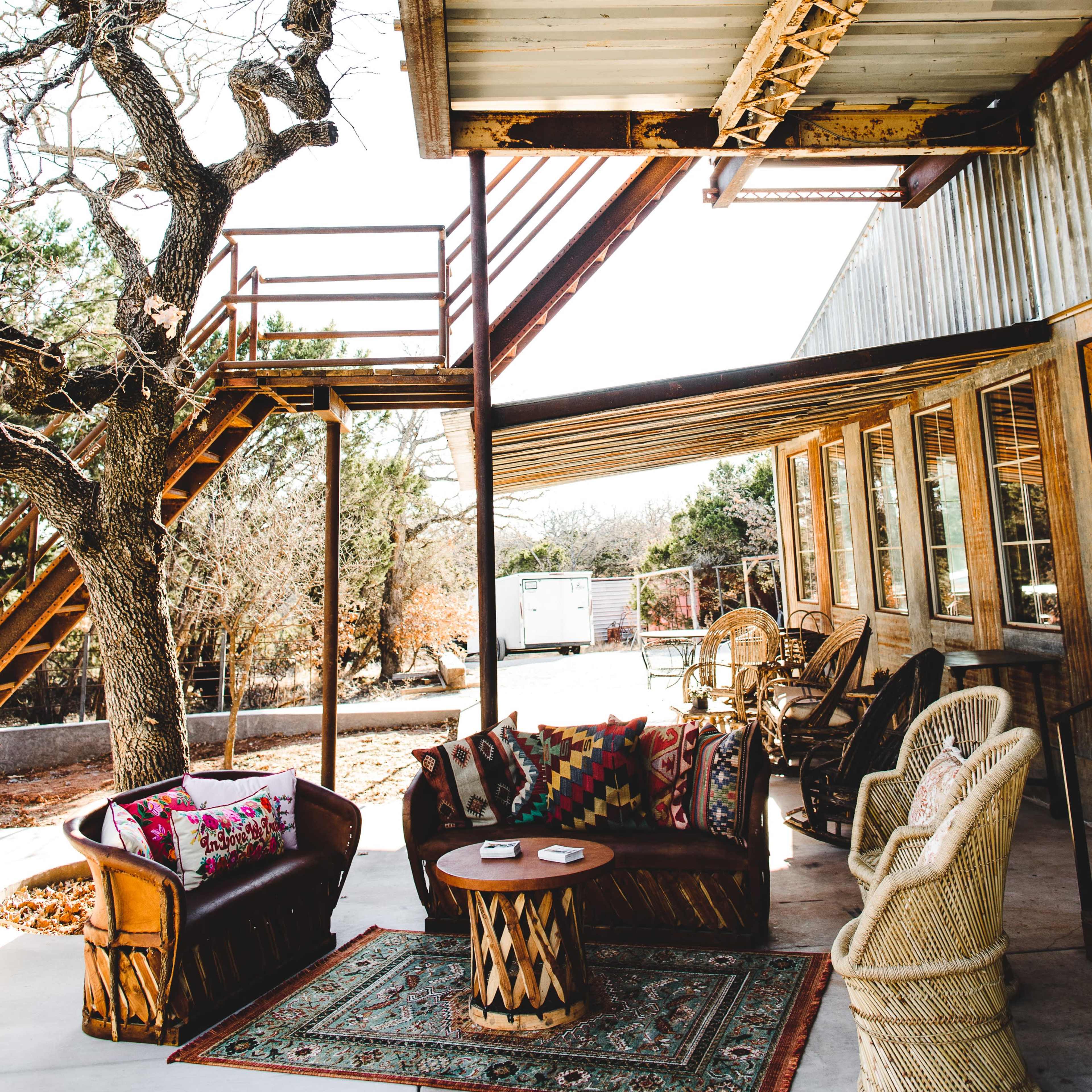 A cozy outdoor seating area features a wooden couch, a round table, and a rug beneath a staircase and a tree.