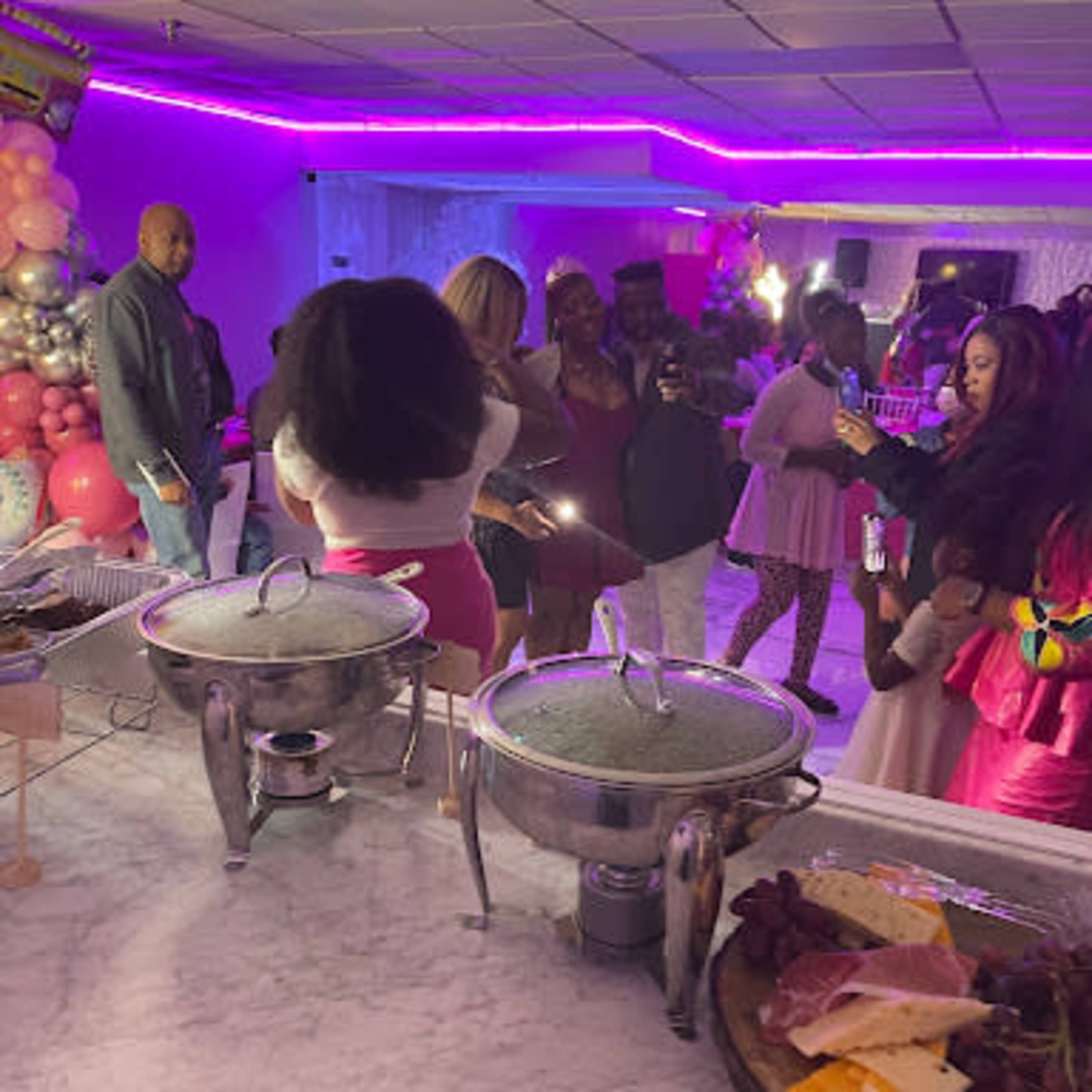 A party scene with guests mingling, a buffet table featuring food, and colorful lighting in the background.