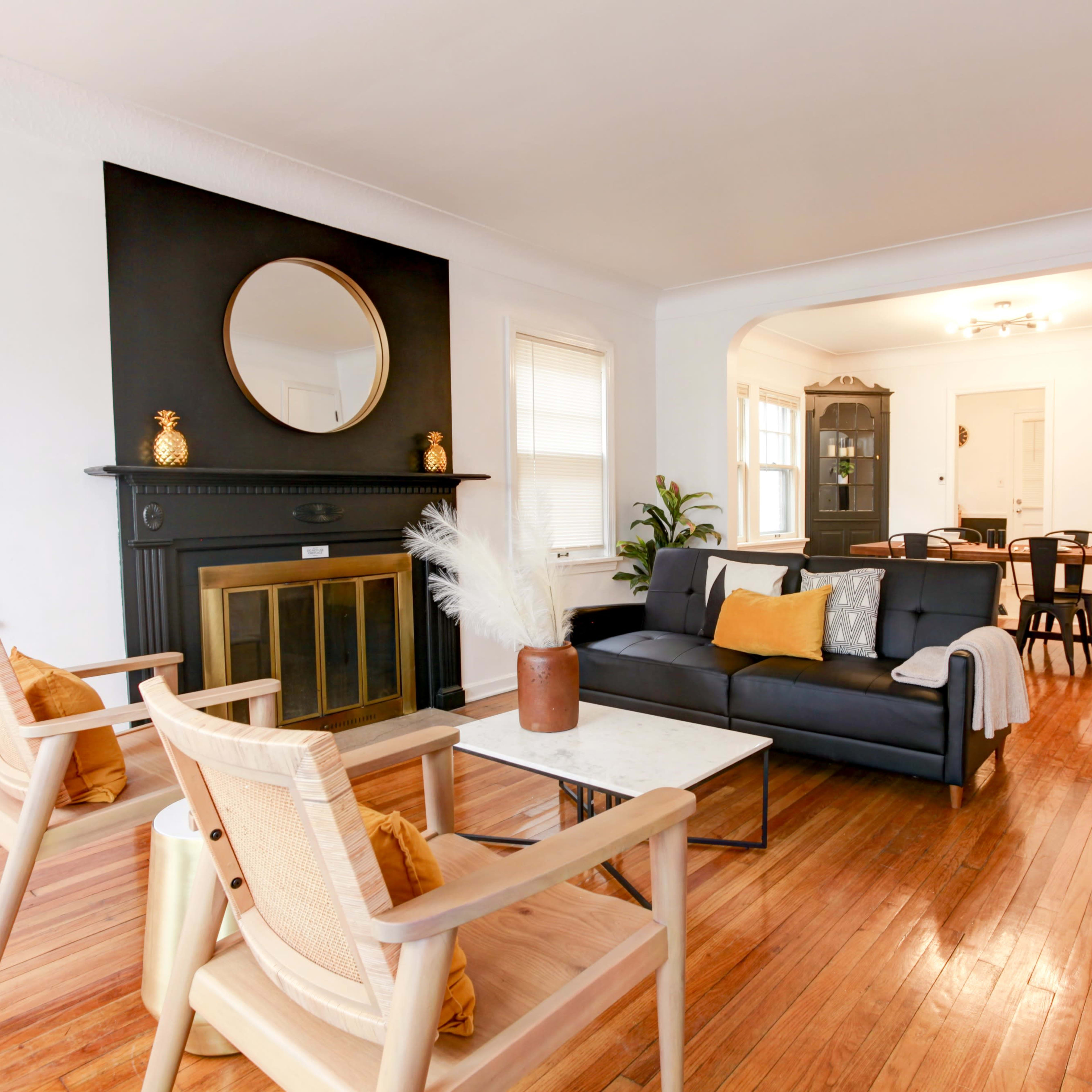 A living room features a black fireplace with a round mirror above it, a sofa with an orange accent pillow, wooden chairs, and a dining area visible in the background.