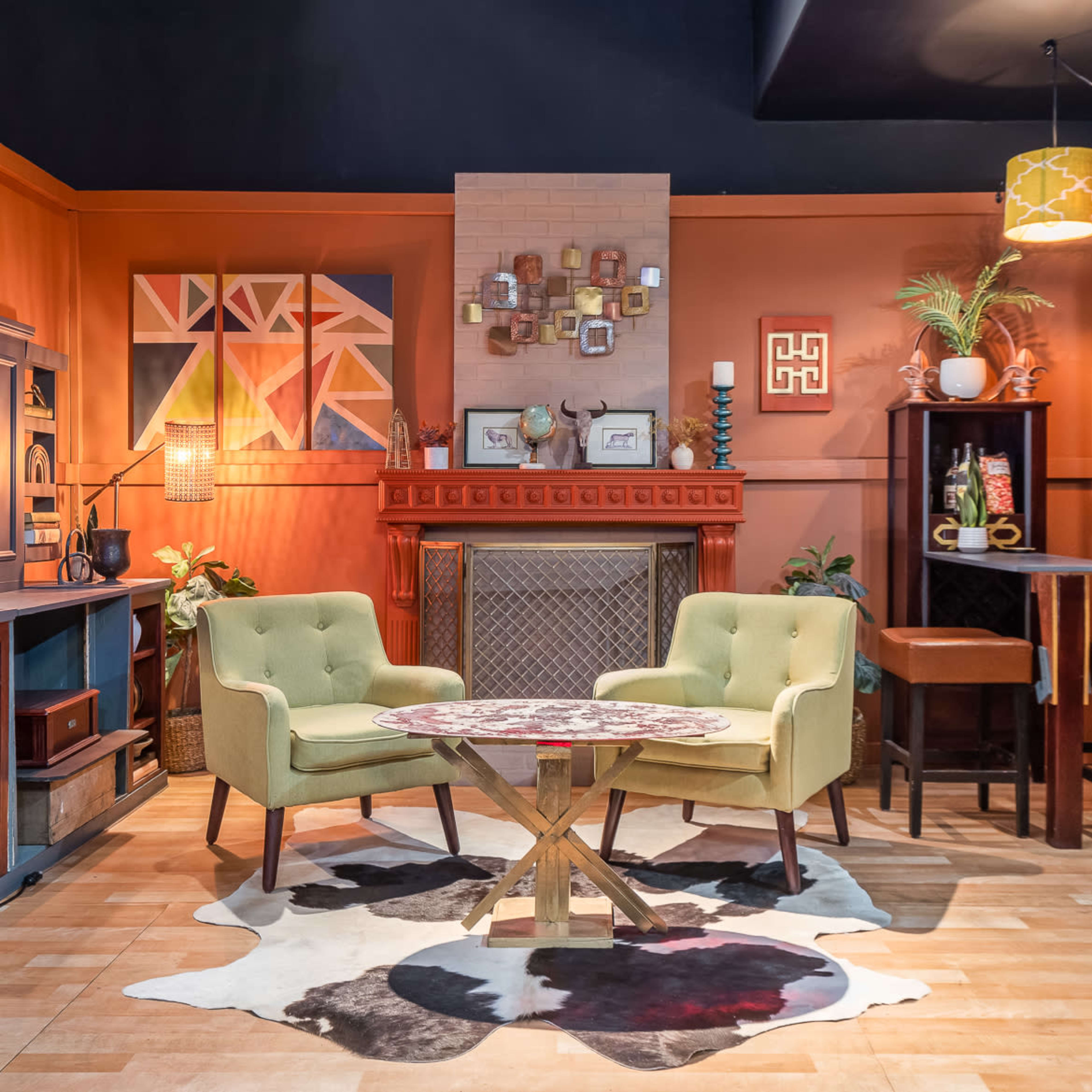 A living room scene features two green armchairs facing a round table, a fireplace with wall art, a shelf unit, and a bar area with stools, surrounded by warm-toned walls and decorative plants.