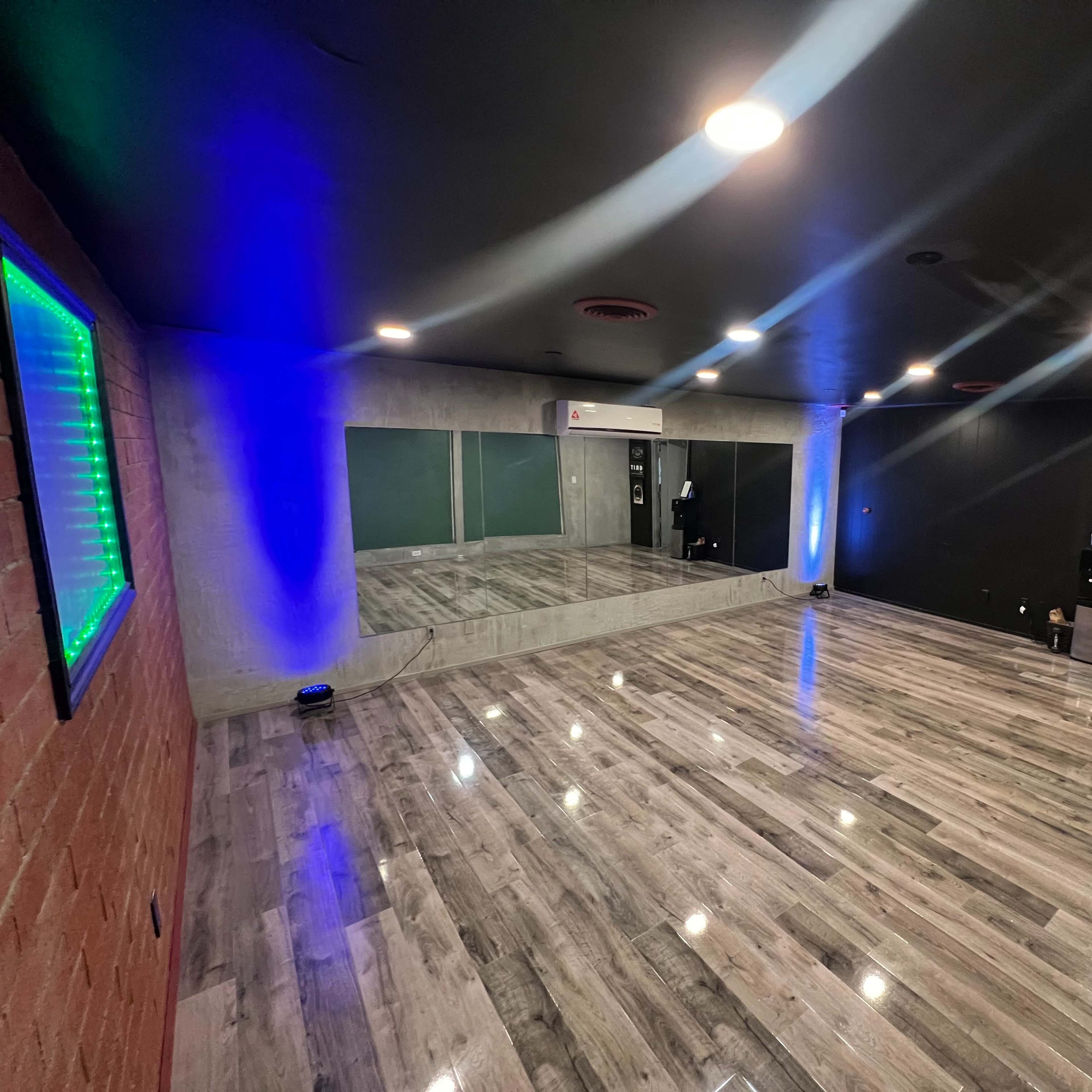 A spacious room with polished wooden floors, a large mirror on one wall, and blue and green lighting accents.