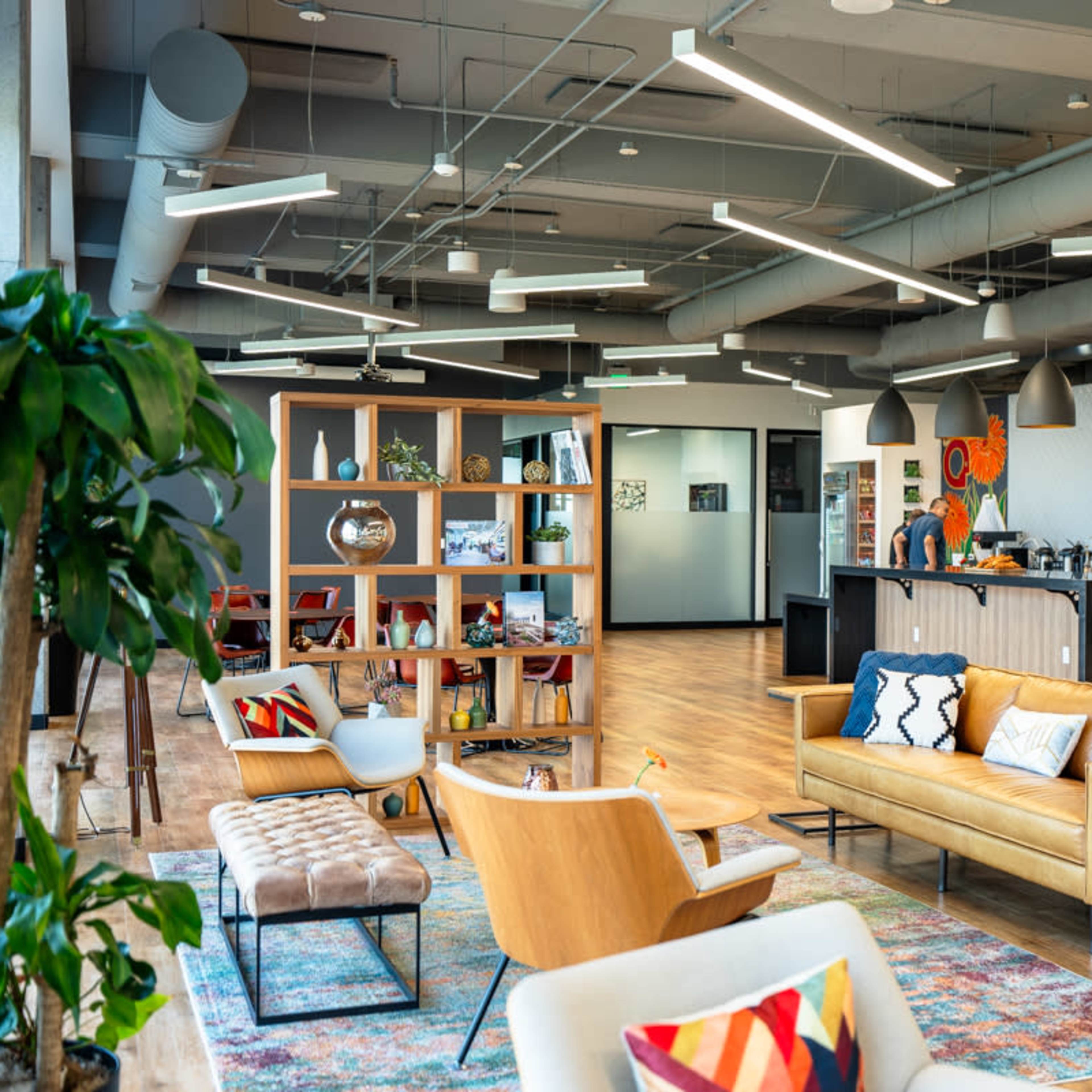 A modern office lounge area with comfortable seating, a bookshelf, and a kitchen counter, featuring wooden floors and ample natural light.