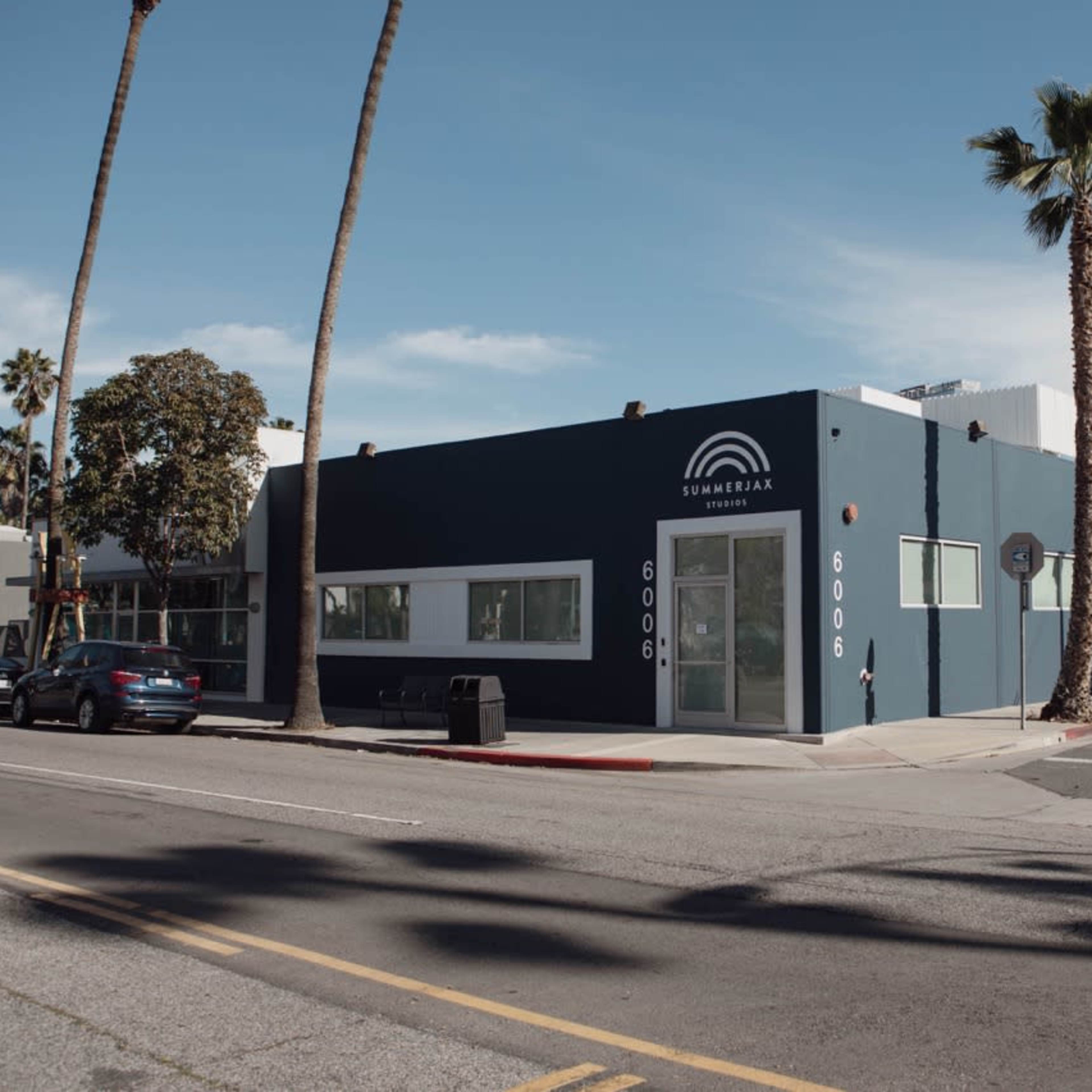 A blue commercial building with large windows stands at the corner of a street, surrounded by palm trees and parked cars.