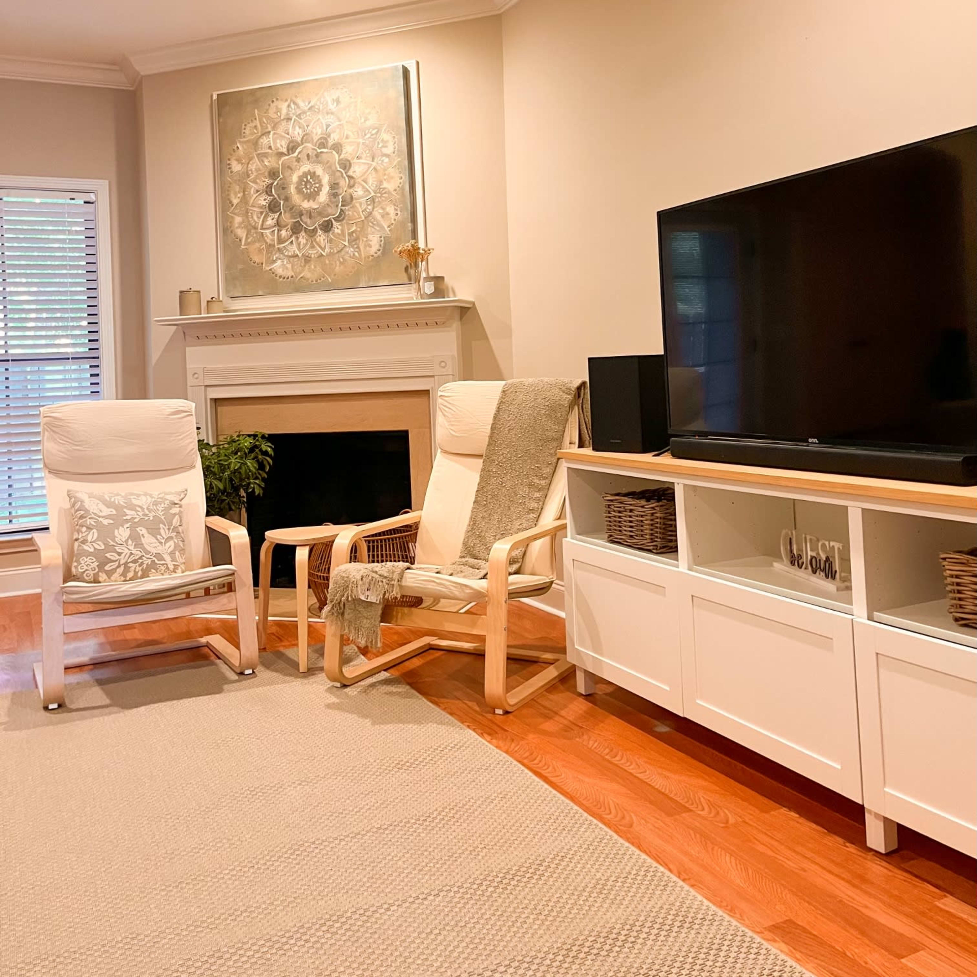 A living room features two wooden chairs, a television on a white cabinet, and a fireplace with a decorative piece above it.