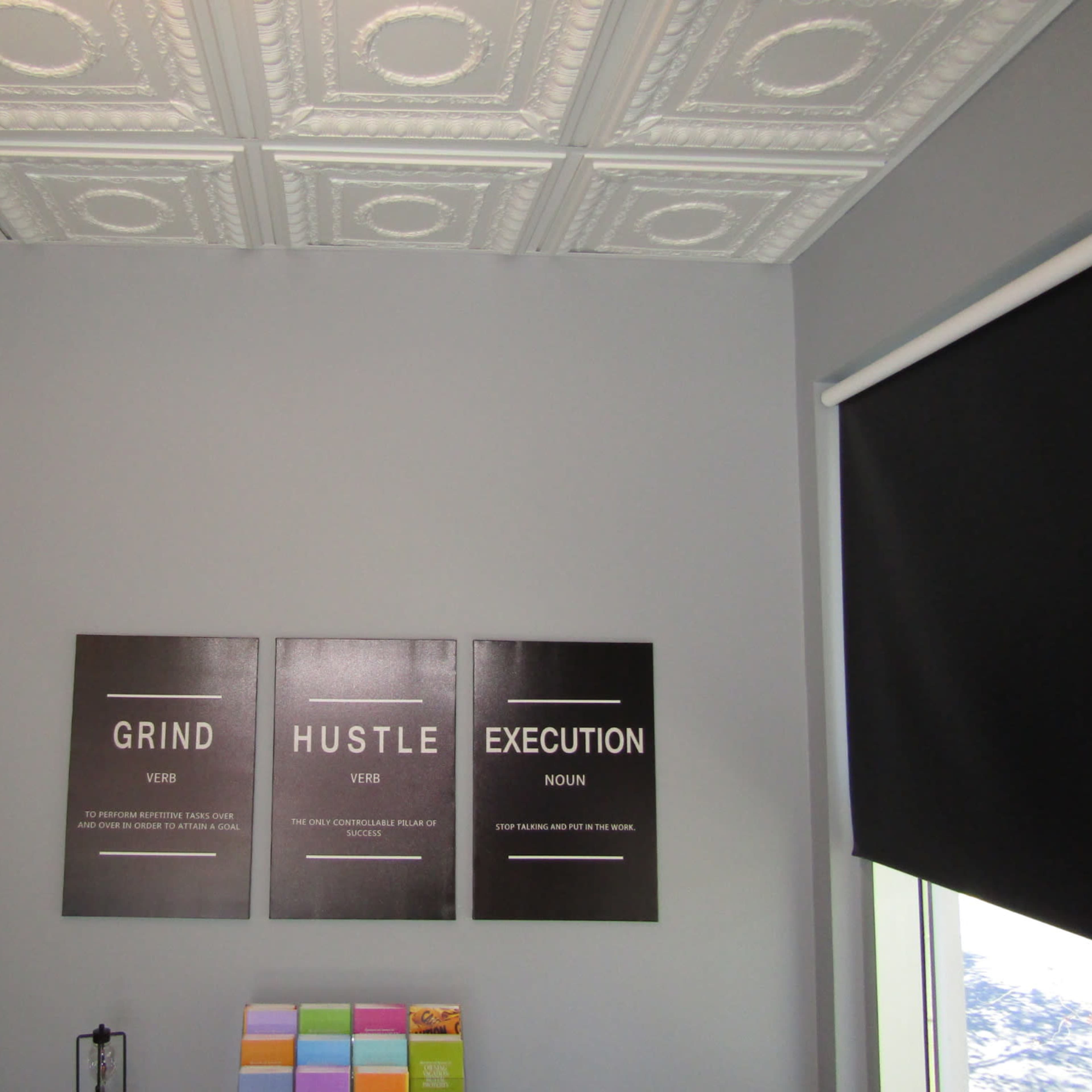 A gray wall displaying three framed signs with the words "GRIND", "HUSTLE", and "EXECUTION" along with their definitions, next to a colorful stack of paper.