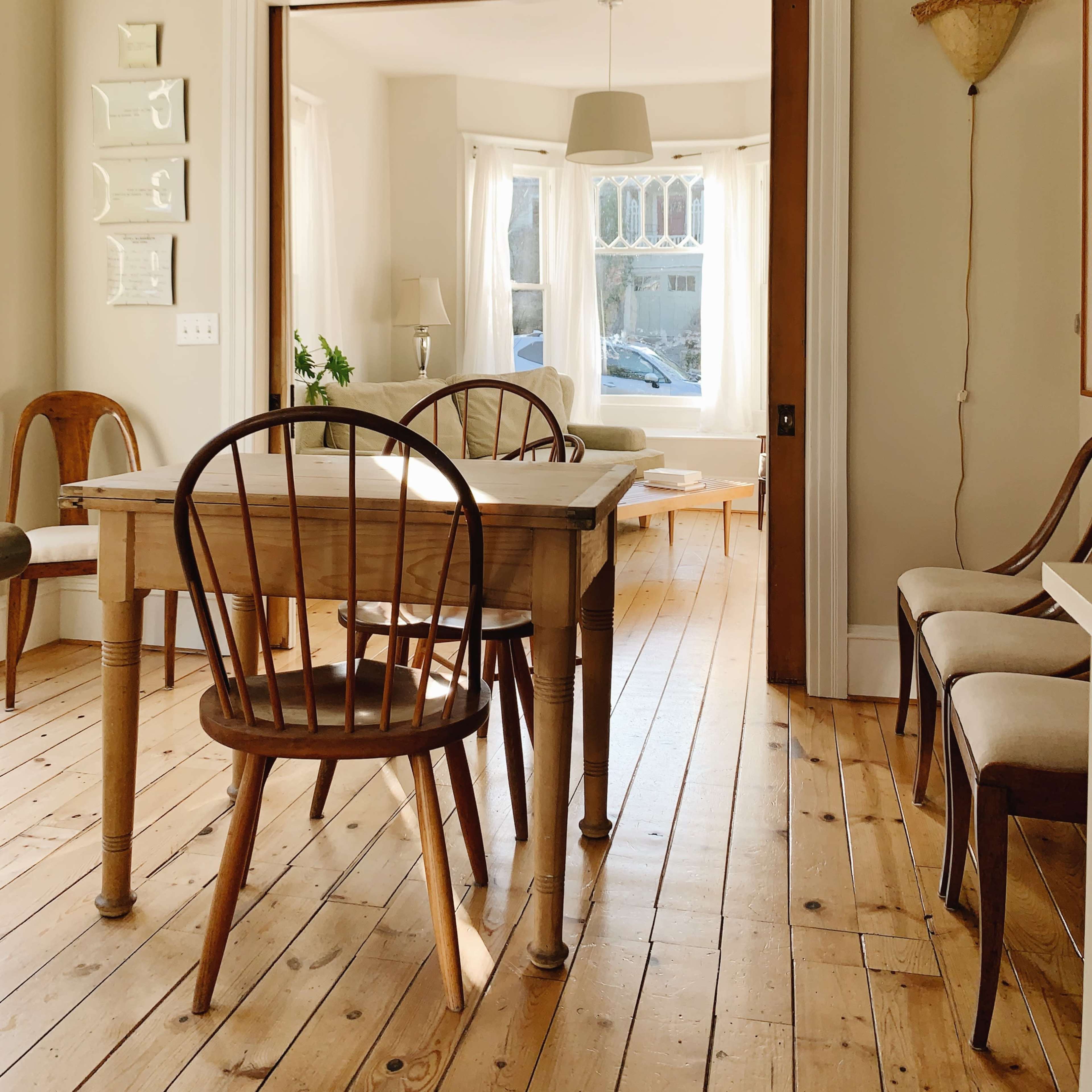 A bright dining area features a wooden table surrounded by chairs, with a view into a sunlit room through an open doorway.