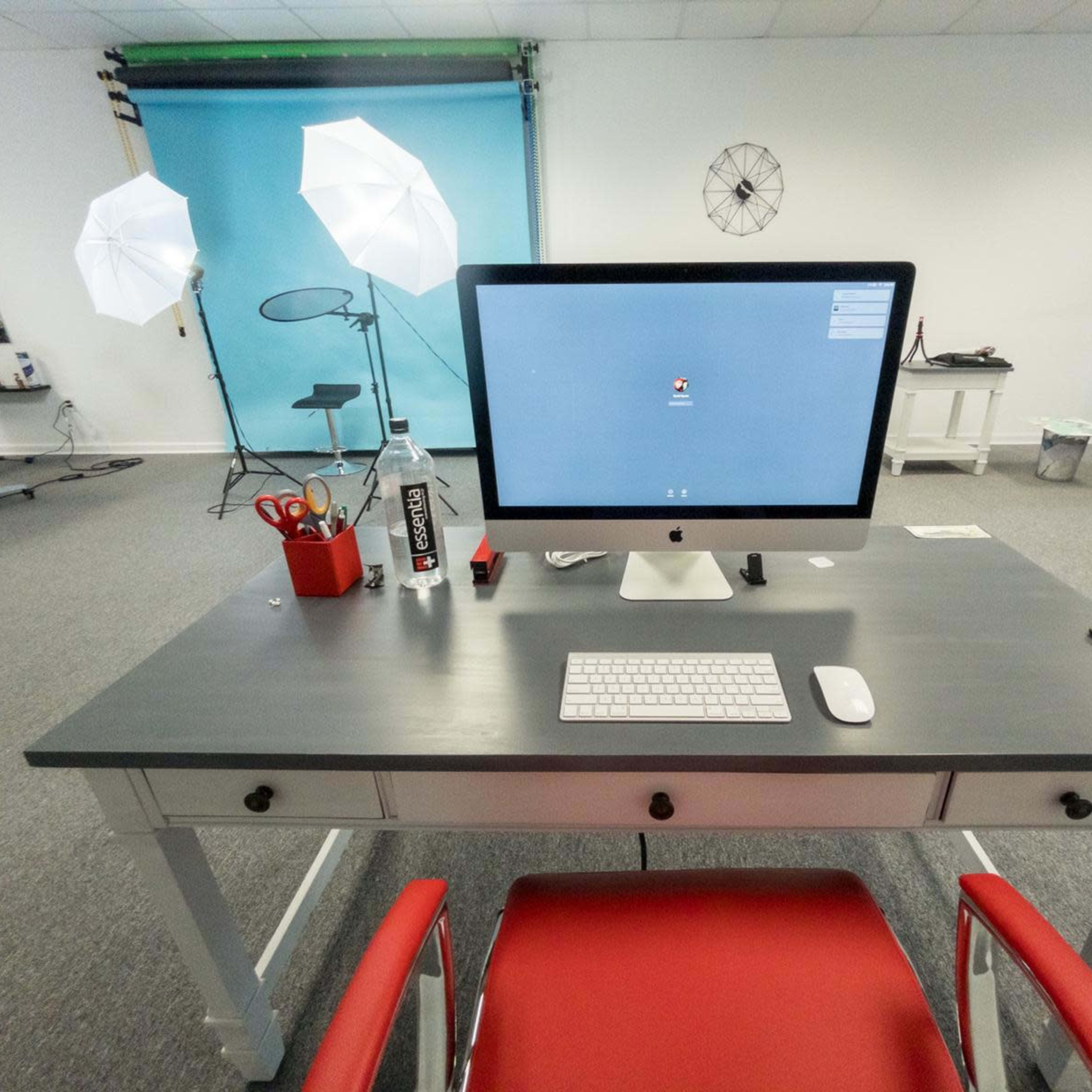 A desk with an iMac computer, keyboard, and various accessories is positioned in a bright studio space featuring photography lighting setups in the background.
